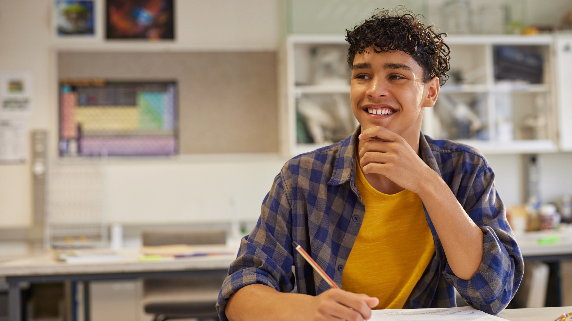 A teenage boy with curly hair smiling and resting his chin on his hand, sitting at a desk with a pencil in hand, in a classroom setting.
