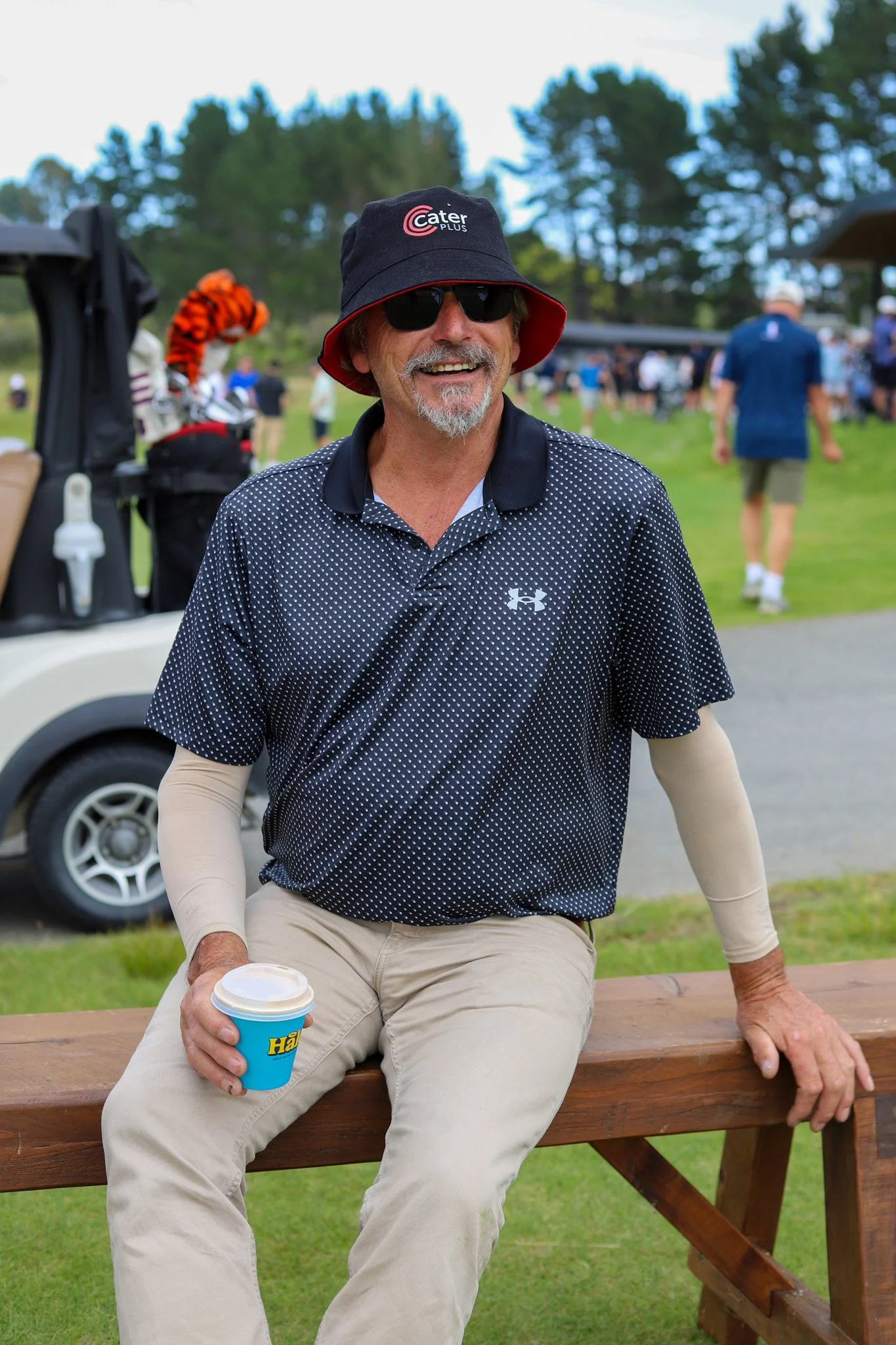 A smiling man sitting on a wooden bench at a golf course, holding a cold drink in a blue cup. He is wearing a black bucket hat with the logo 'Cater Plus,' sunglasses, a black Under Armour golf shirt, and beige pants. There are people and a golf cart in the background on a grassy area with trees.