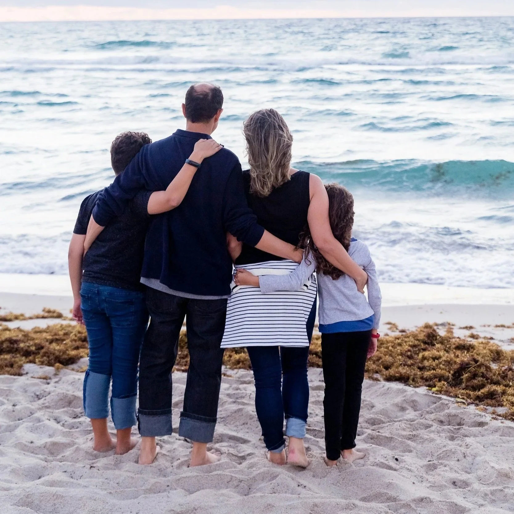 Family standing on a beach facing the ocean, with their arms around each other.