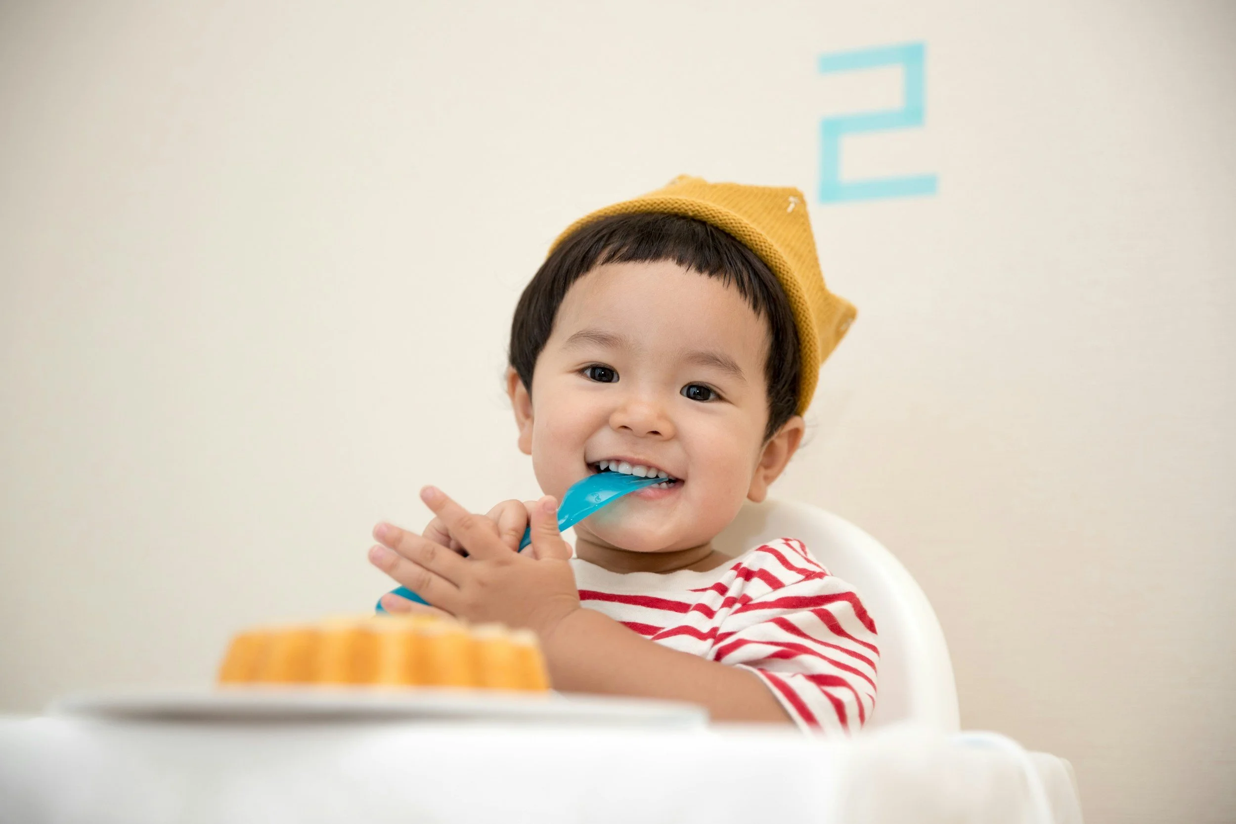 A young boy with dark hair, wearing a yellow hat and a red and white striped shirt, happily eating with a blue spoon at a high chair.