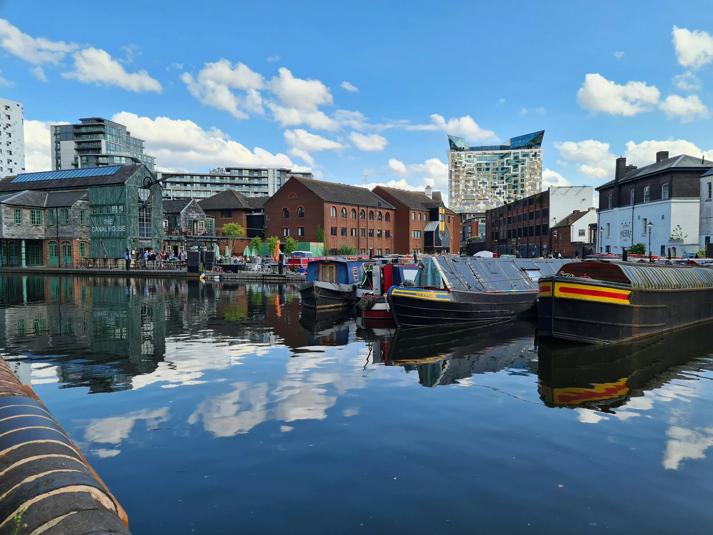 Photo of Birmingham City Centre taken from a canel toe path, looking towards the Cube. In the foreground of the photo are several moored narrow boats and The Canal House building.