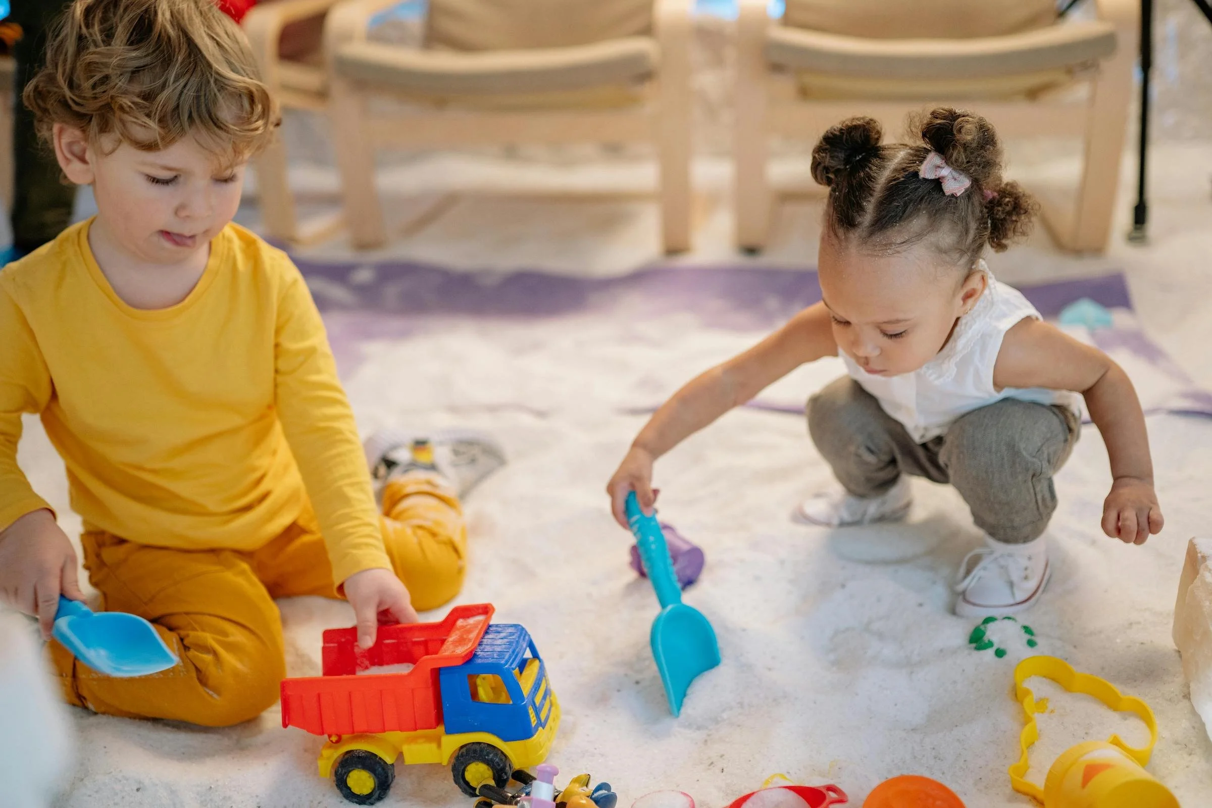 Two young children, a boy with curly blond hair and a girl with curly hair styled in two buns, are playing in a sandbox. They are surrounded by colorful toys, including shovels, buckets, and molds.