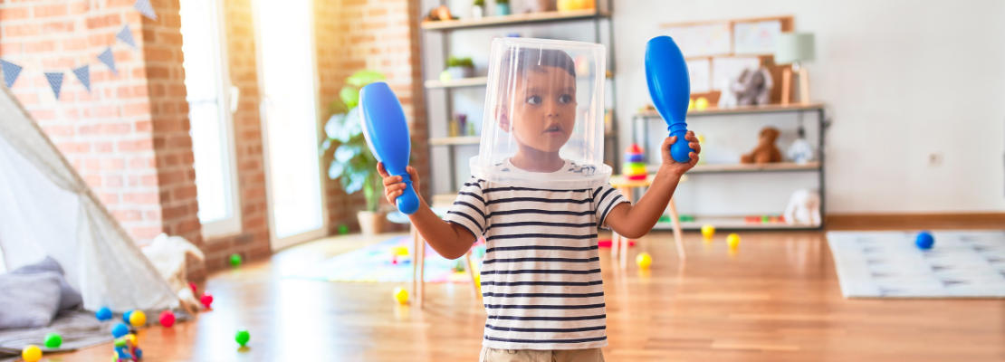 Photo of a young child in a nursery setting with a clear plastic bucket on their head and holding up two blue plastic skittles.