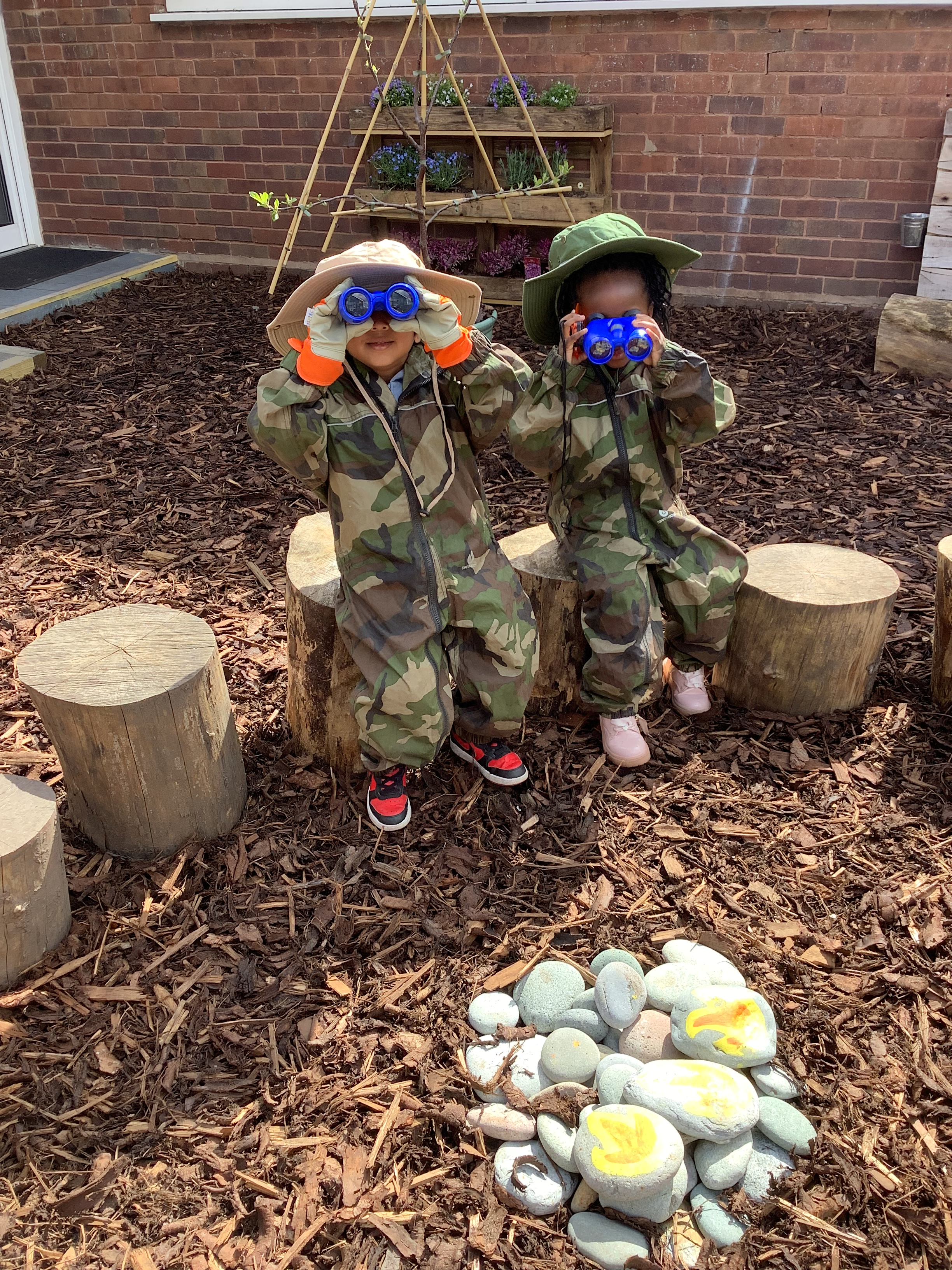 Photo of two young children in camouflage playsuits and floppy summer hats playing in The Nest. They are both holding bright blue binoculars up to their eyes and looking at the camera.