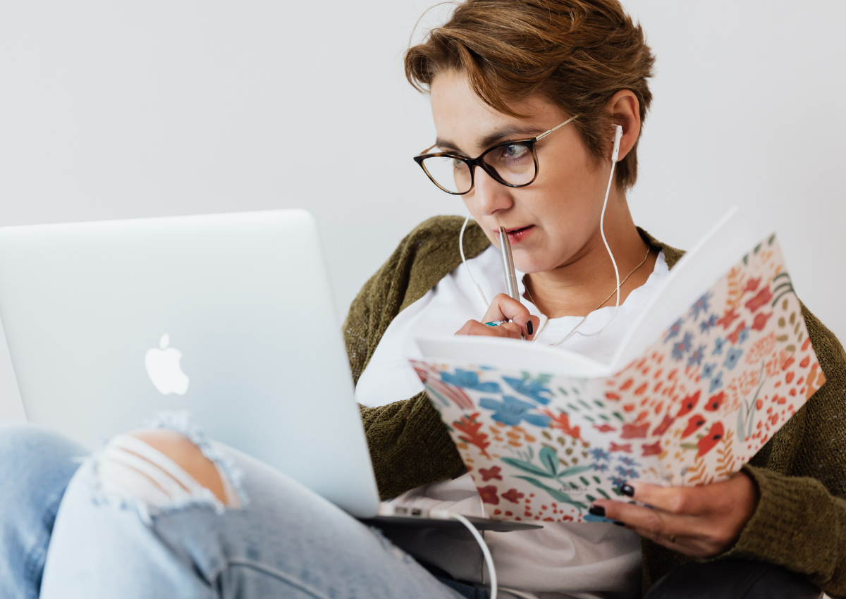 A white, fem-presenting person with short hair and wearing glasses, watching a webinar on their laptop while taking notes in a colourful, floral notebook.