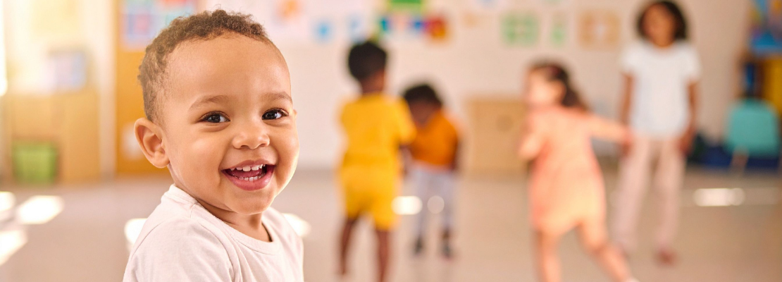 Close-up photo of a young child in an early years setting smiling at the camera. Other children can be seen playing and dancing out-of-focus in the background.