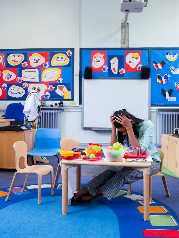 Photo of a stressed Nursery Headteacher in an empty classroom. She is sitting at the toy table with her head in her hands.