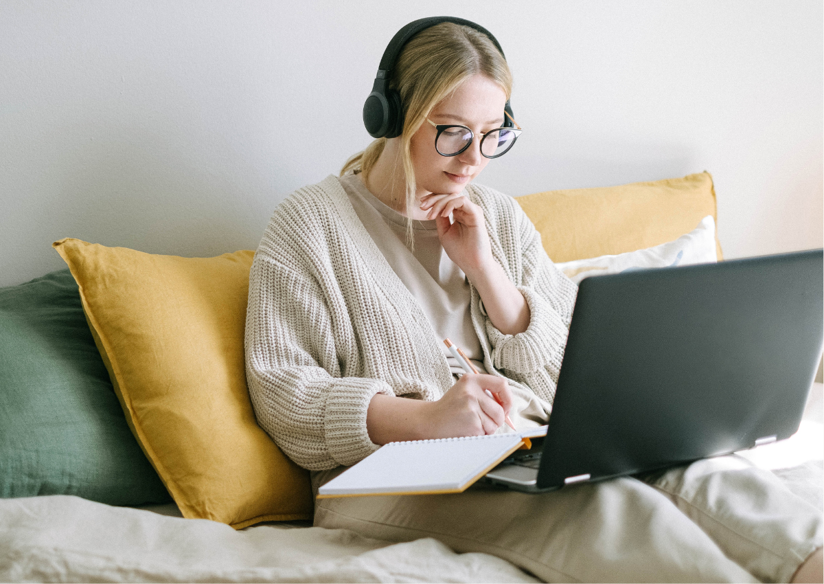 A person sat on a comfy couch with their laptop and a notebook listening to a webinar recording and taking notes.
