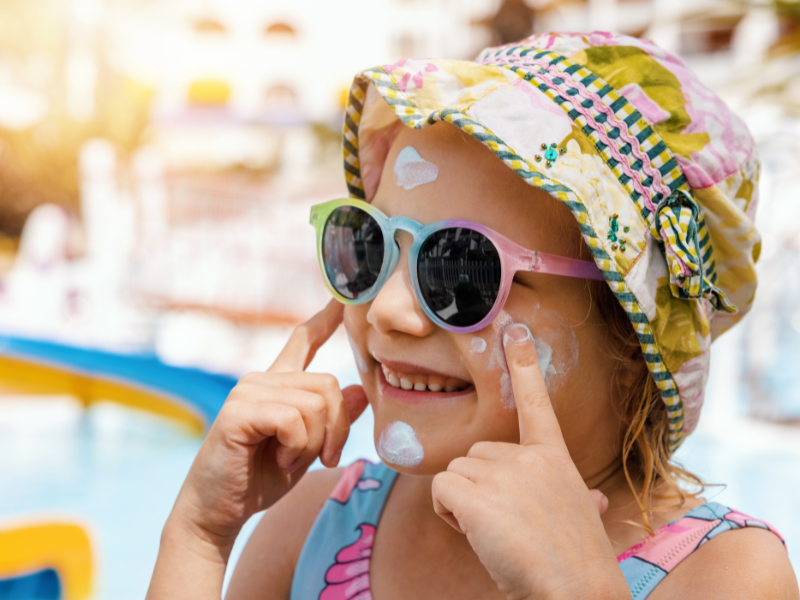 Close-up photo of a young child wearing a wide-brimmed sun hat and sunglasses, putting sunscreen on their face.