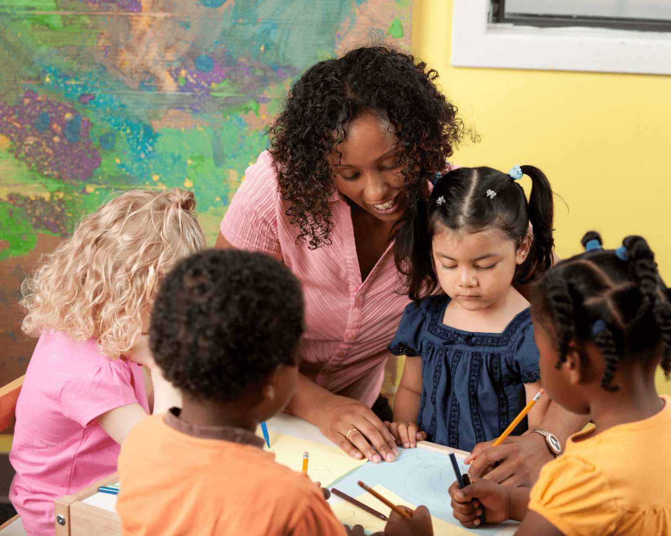 A light skinned black woman wearing a pink shirt is helping a little girl, who has her hair in bunches and is wearing a blue short sleeved dress, with her handwriting. Two other children watch on from the foreground, slightly out of focus