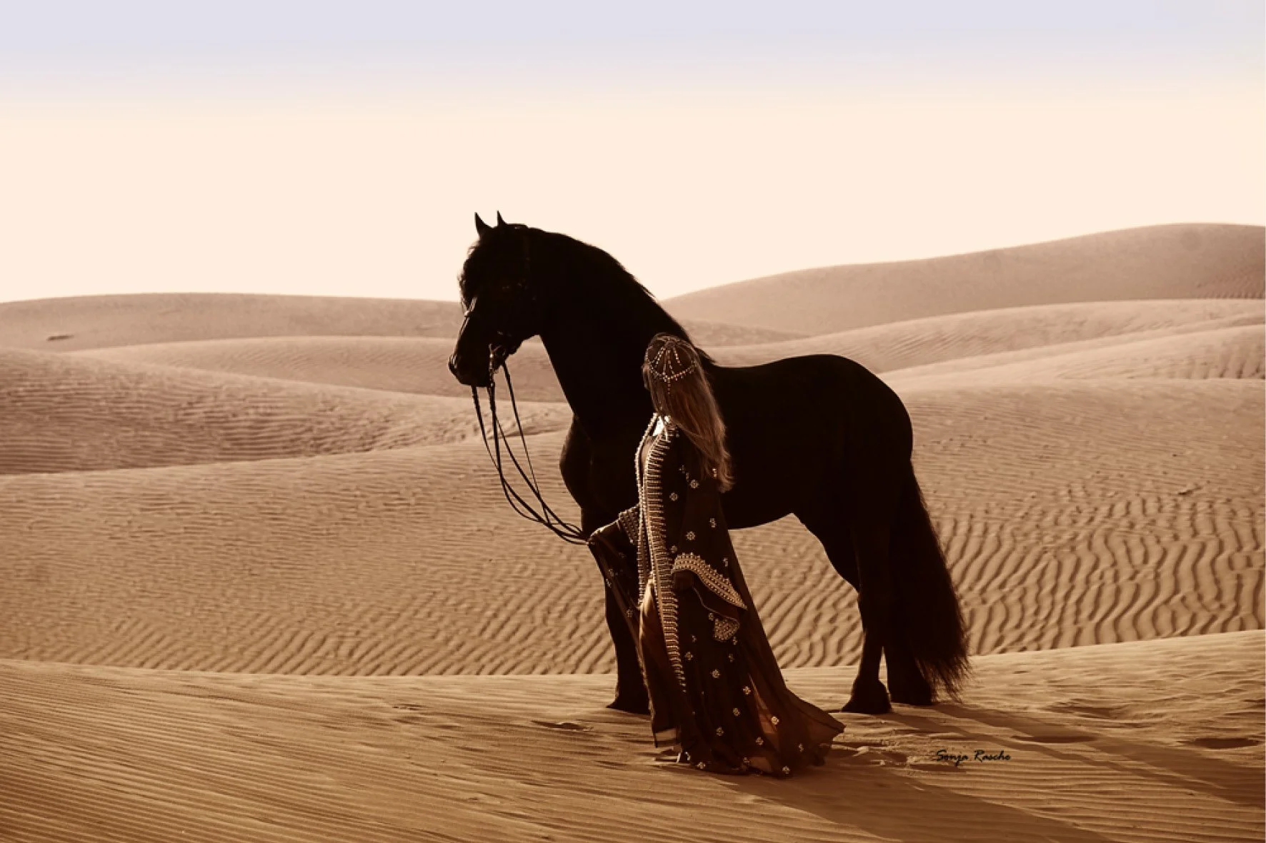A person in traditional clothing standing beside a black horse in a desert landscape.