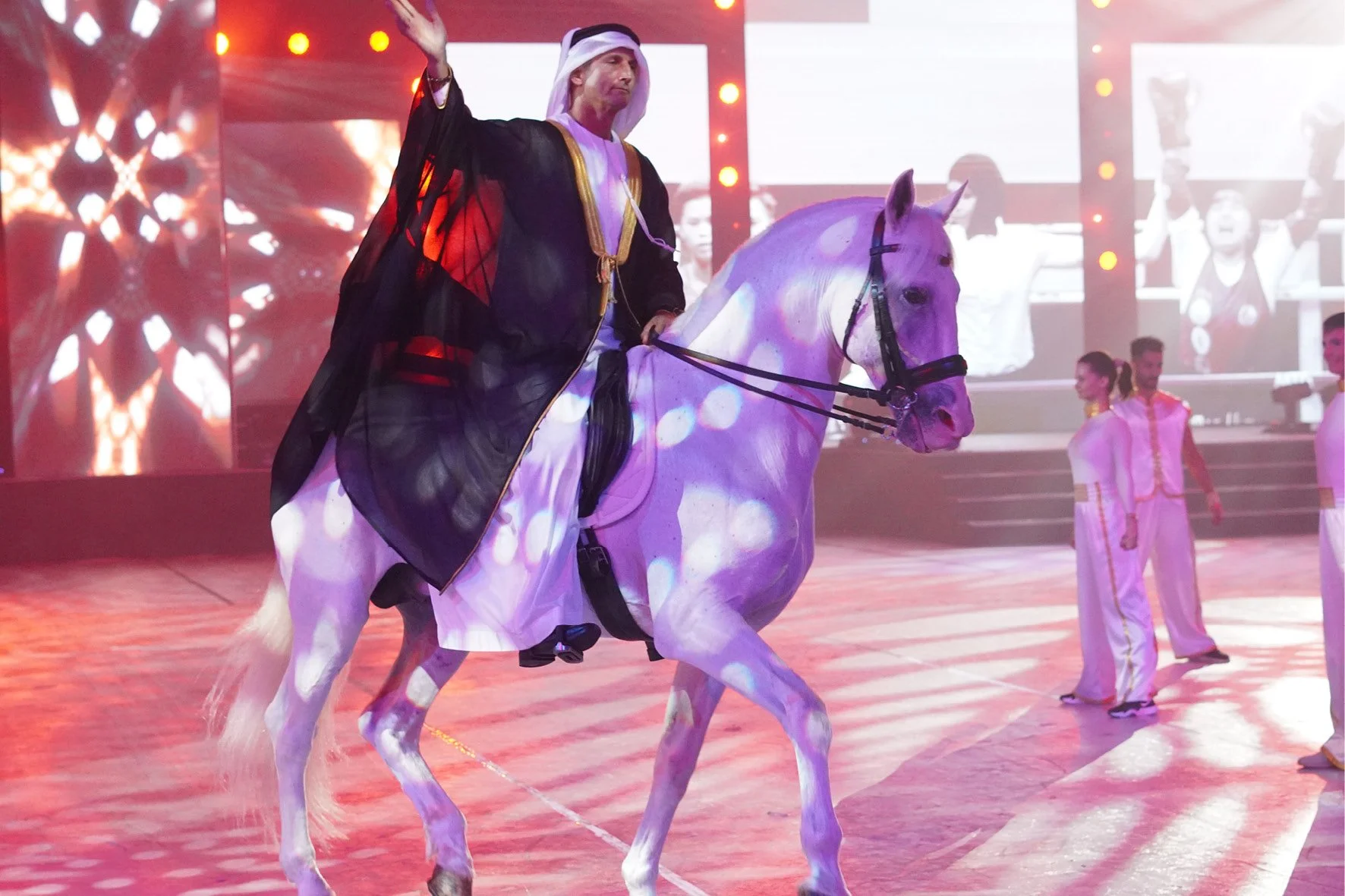 Man in traditional outfit riding a white horse in a lit indoor arena with performers in background.