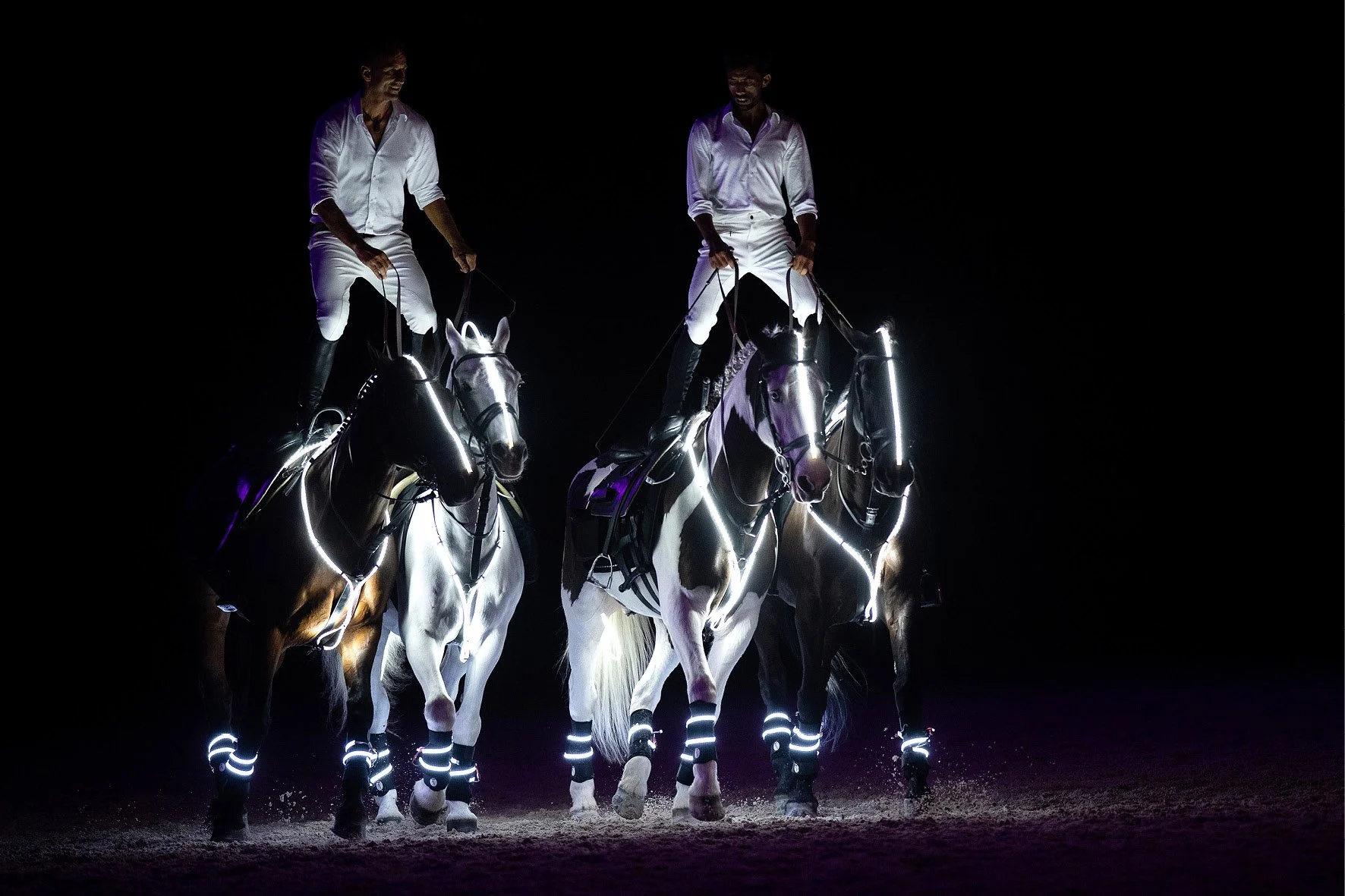 Two riders in white attire on horses with glowing LED decorations against a dark background.