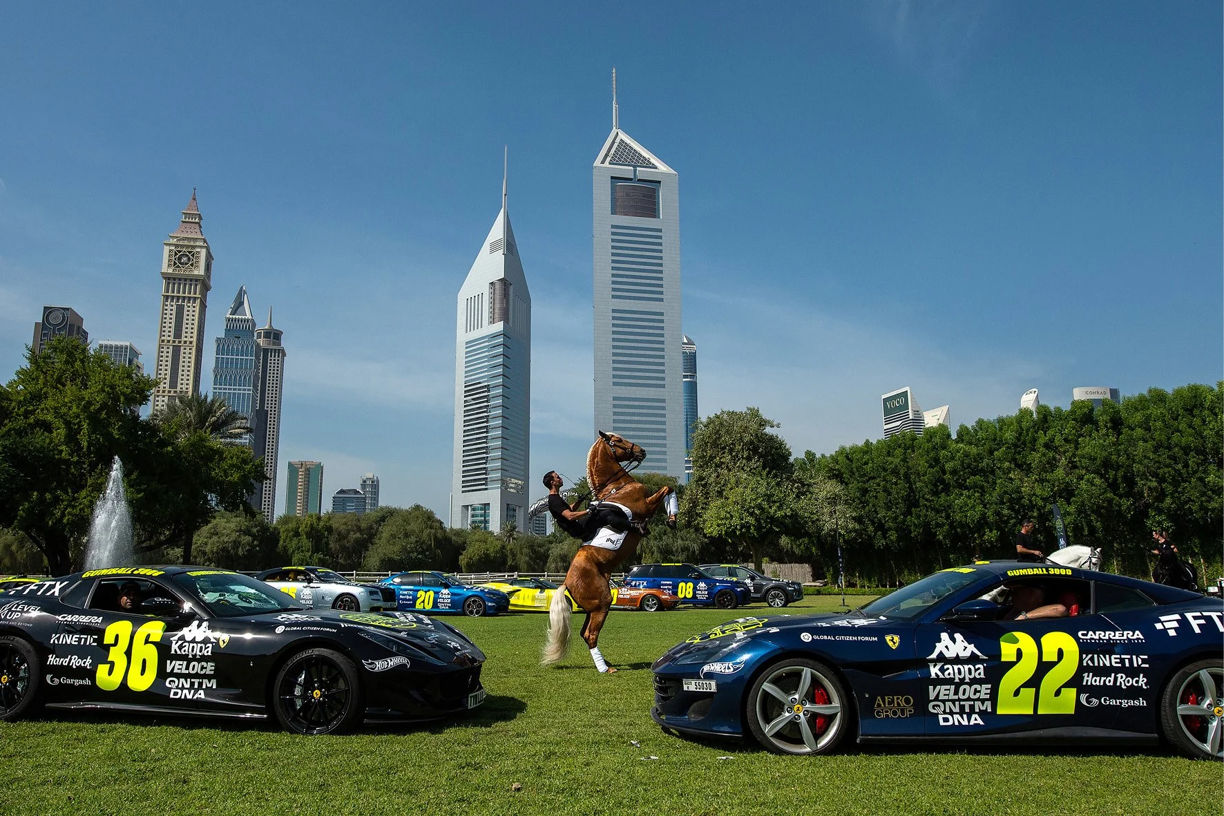 Cars with racing numbers parked on grass, skyscrapers in background, horse and rider performing trick in the middle.