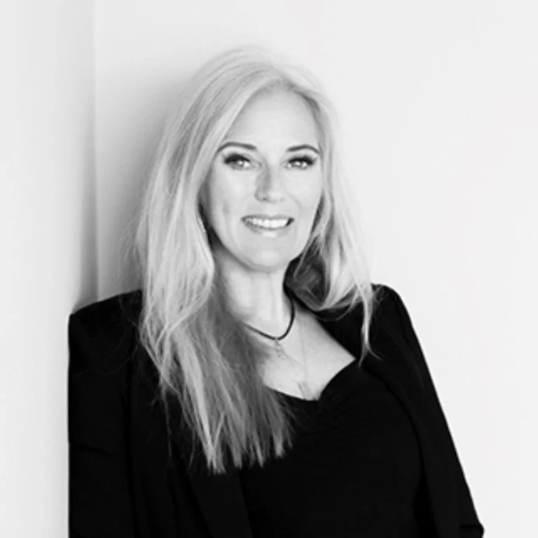 Et sort-hvid portræt af en smilende blond kvinde med langt hår, iført sort tøj, stående mod en lys væg.
