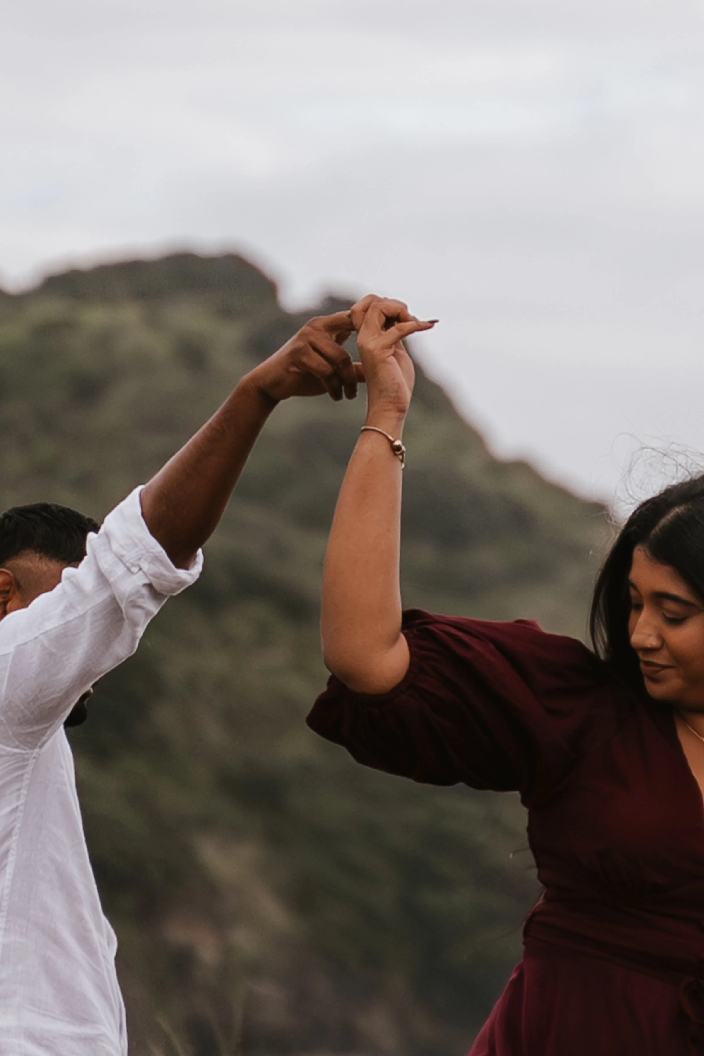 Two people holding hands with interlocked fingers, one wearing a white shirt and the other a maroon dress, against a cloudy sky and mountains in the background.