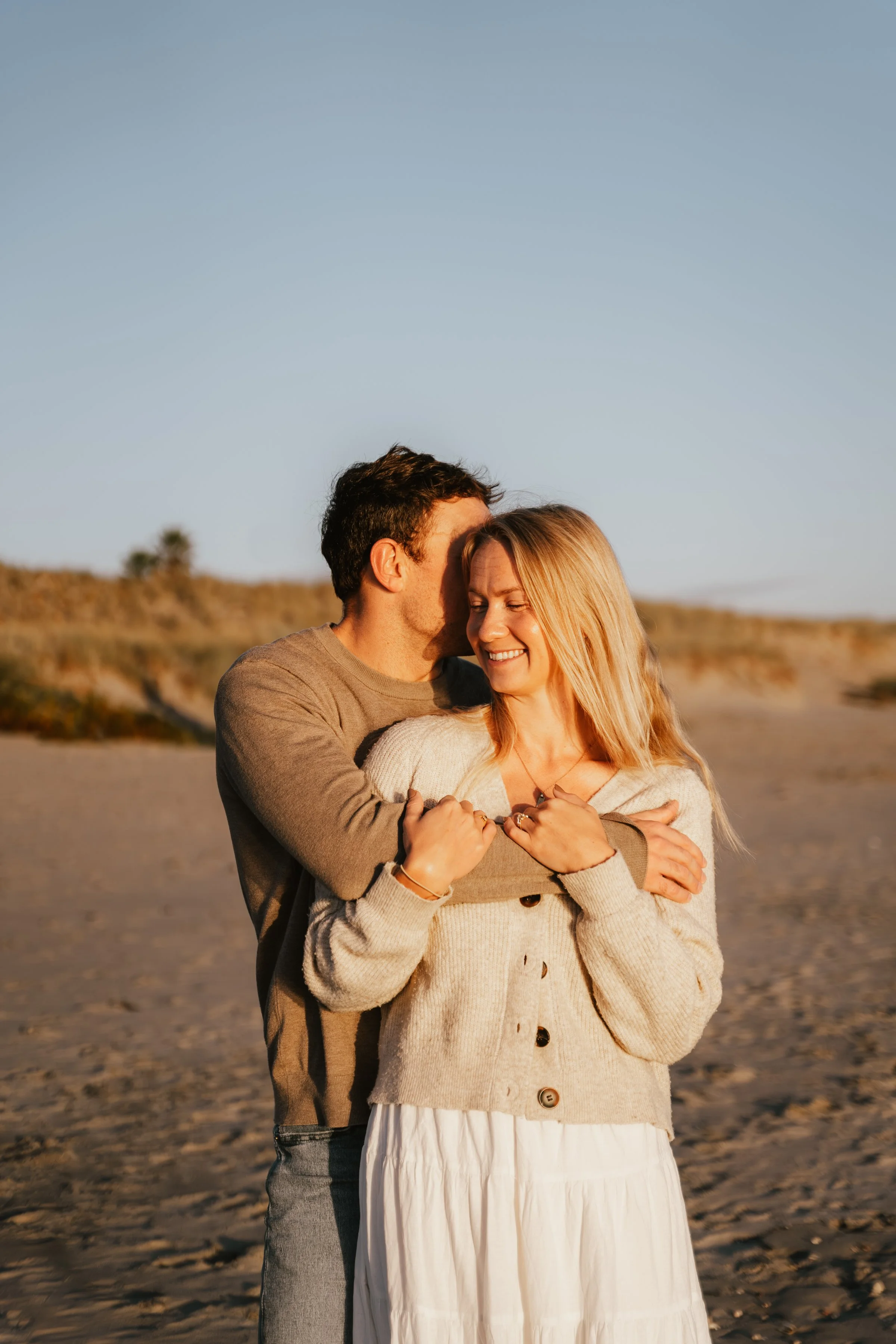 A couple embracing and smiling on a beach during sunset, with a landscape in the background.