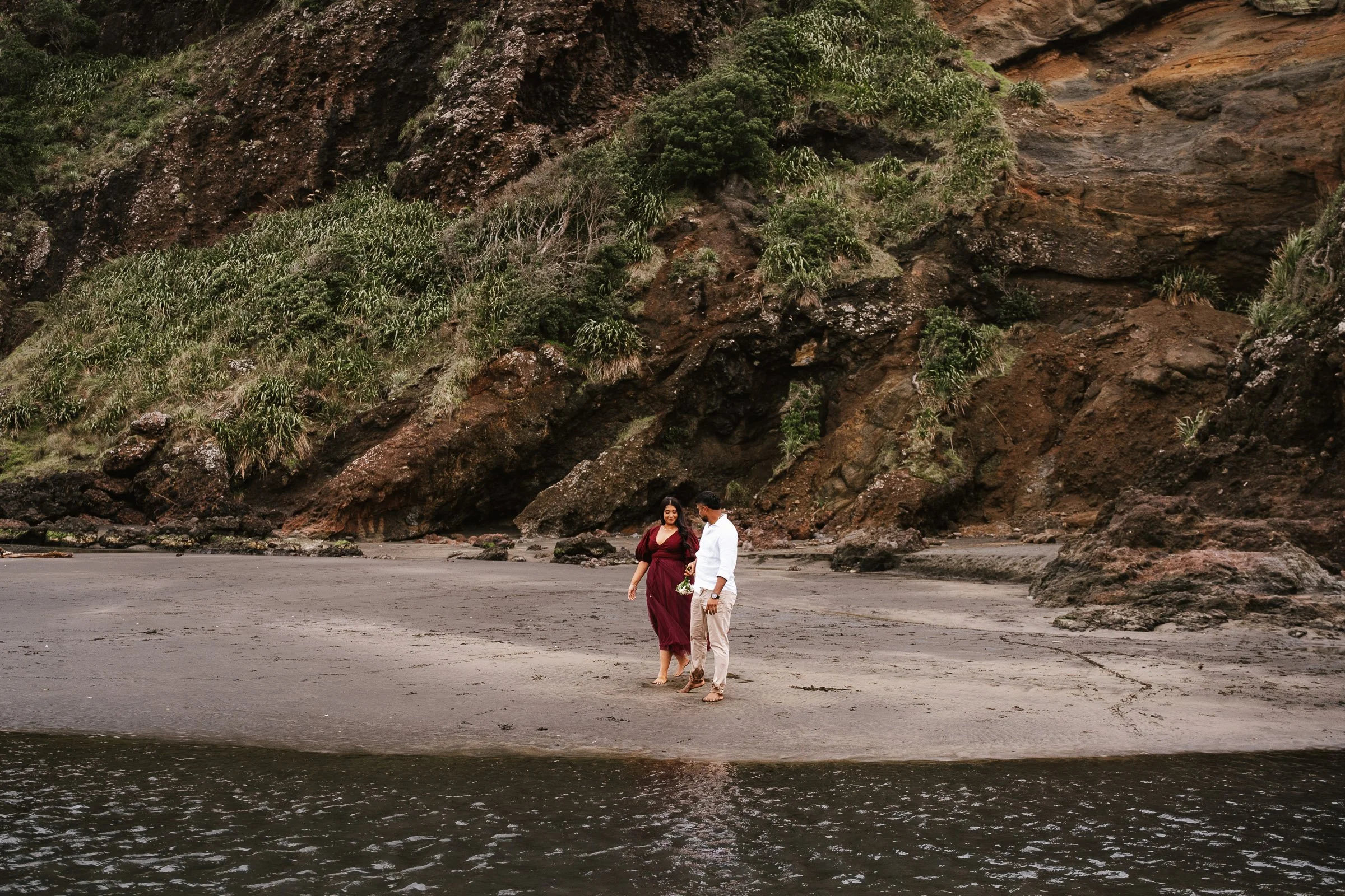 A couple walks hand in hand along a sandy beach near rocky cliffs with green vegetation at the top, during daytime.