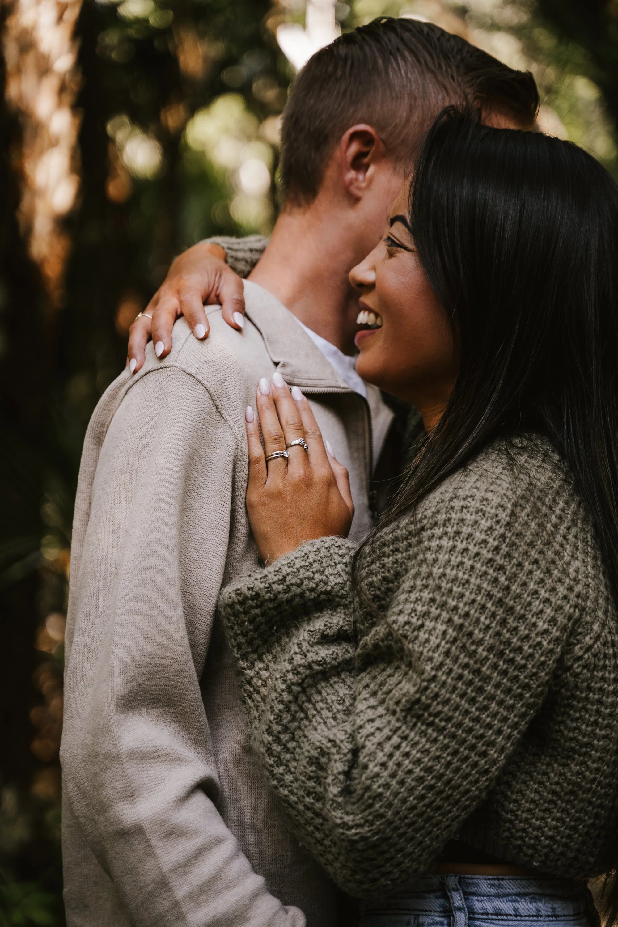 Close-up photograph of a same-sex couple sharing an intimate hug outdoors. The woman with long black hair is smiling with her eyes closed, her hand resting on the man's shoulder, showing multiple rings. The man has short hair and is facing her, with part of his face visible. The background is blurred with natural greenery and warm sunlight.