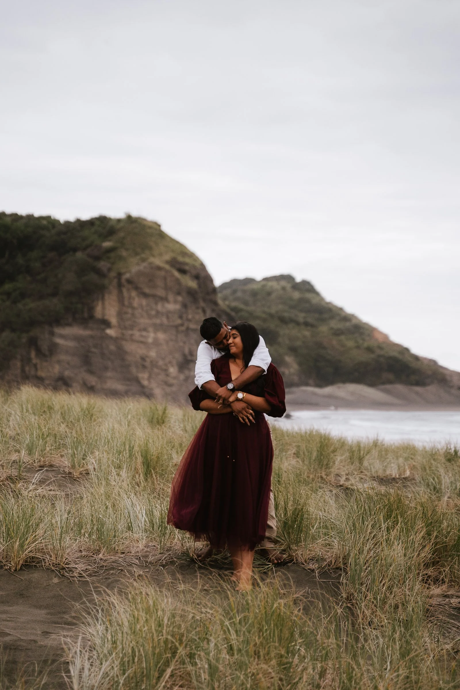A couple embraces on a grassy beach with cliffs in the background, overcast sky.