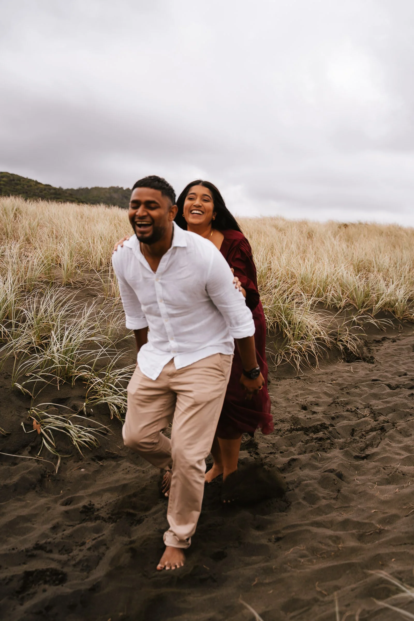 A man and a woman are joyful as they walk barefoot on dark sandy terrain through a grassy field. The man in a white shirt and beige pants leads, while the woman in a maroon dress follows, smiling. The landscape is open with a cloudy sky and distant hills.