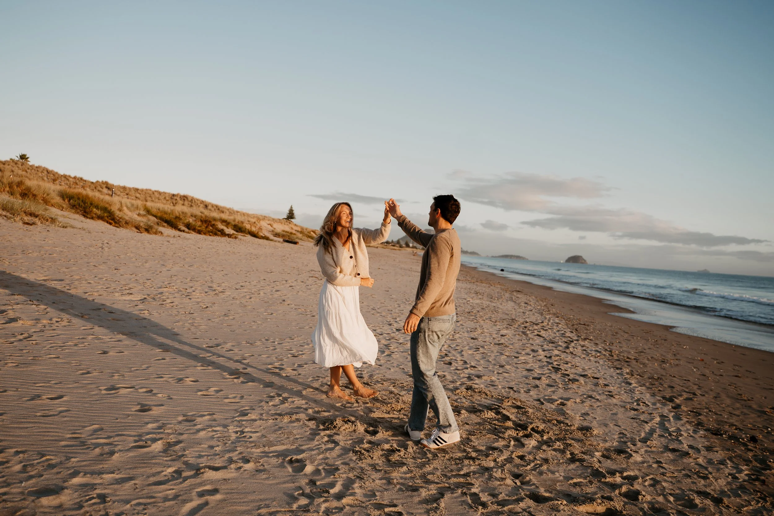 A couple dancing on the beach at sunset, holding hands and smiling.