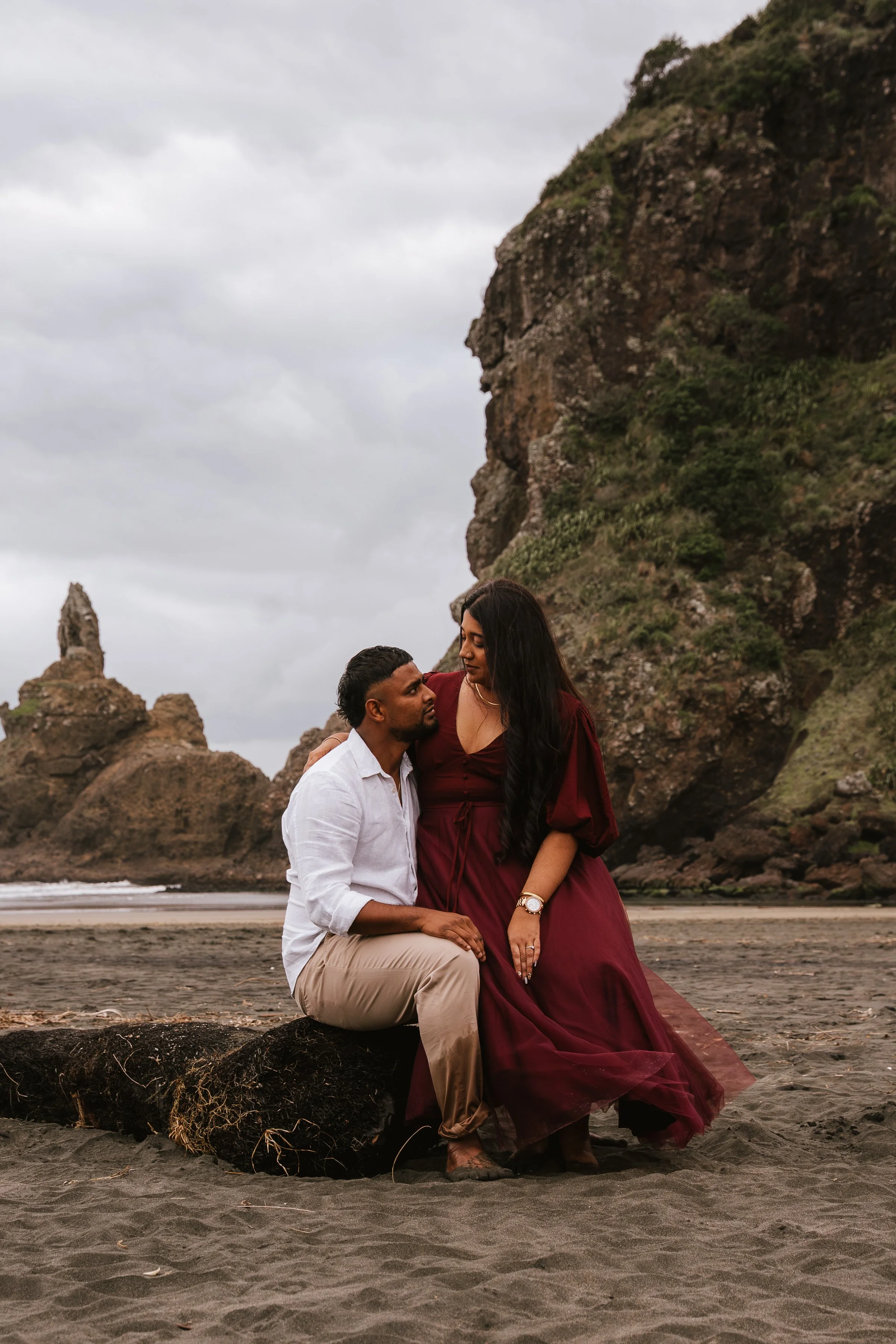 A couple sitting on a large log on a beach, with rocky cliffs and cloudy sky in the background. The woman is dressed in a long burgundy dress, and the man wears a white shirt and beige pants.