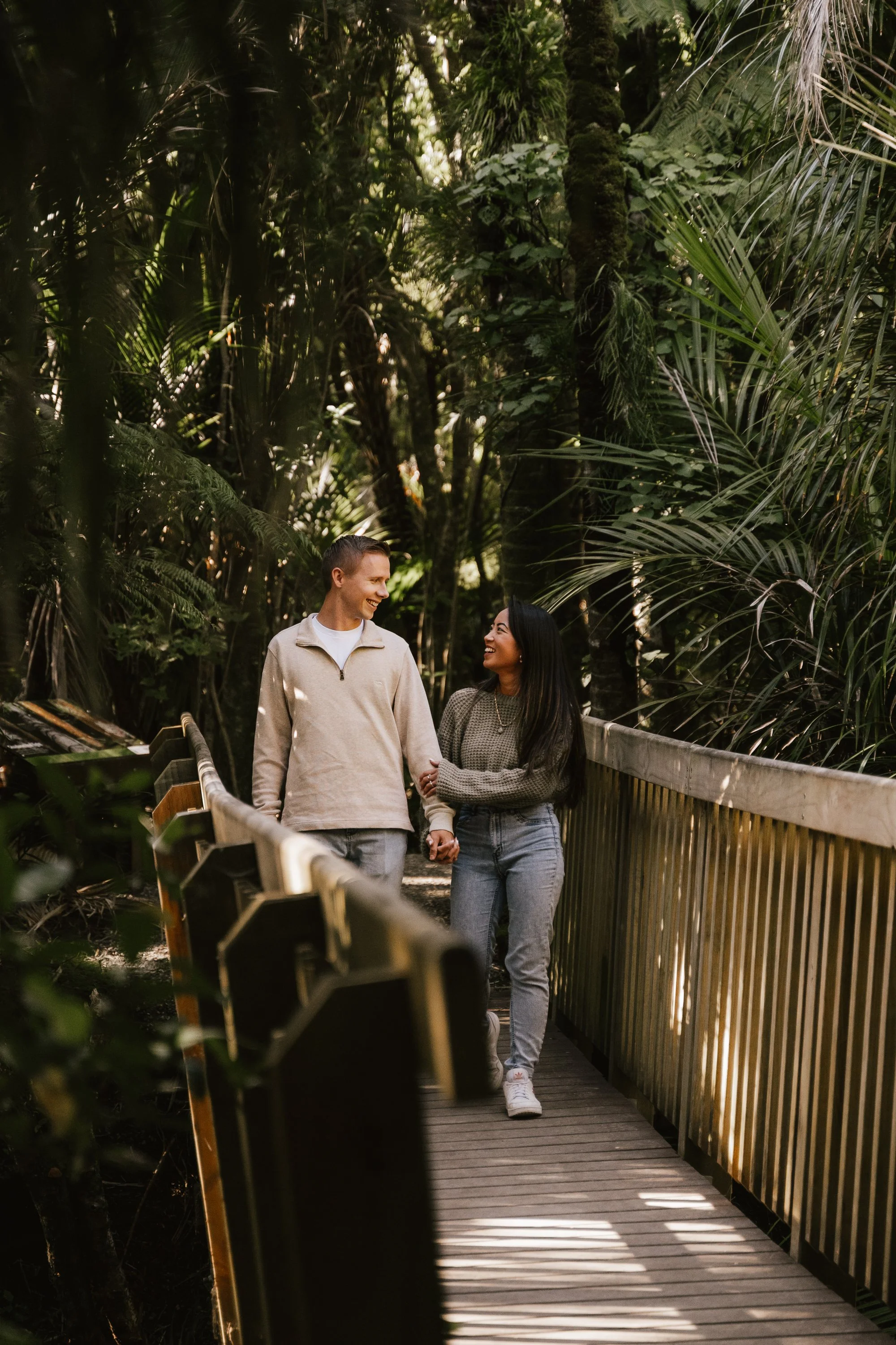A couple walking hand-in-hand on a wooden bridge in a lush green forest, smiling and looking at each other.