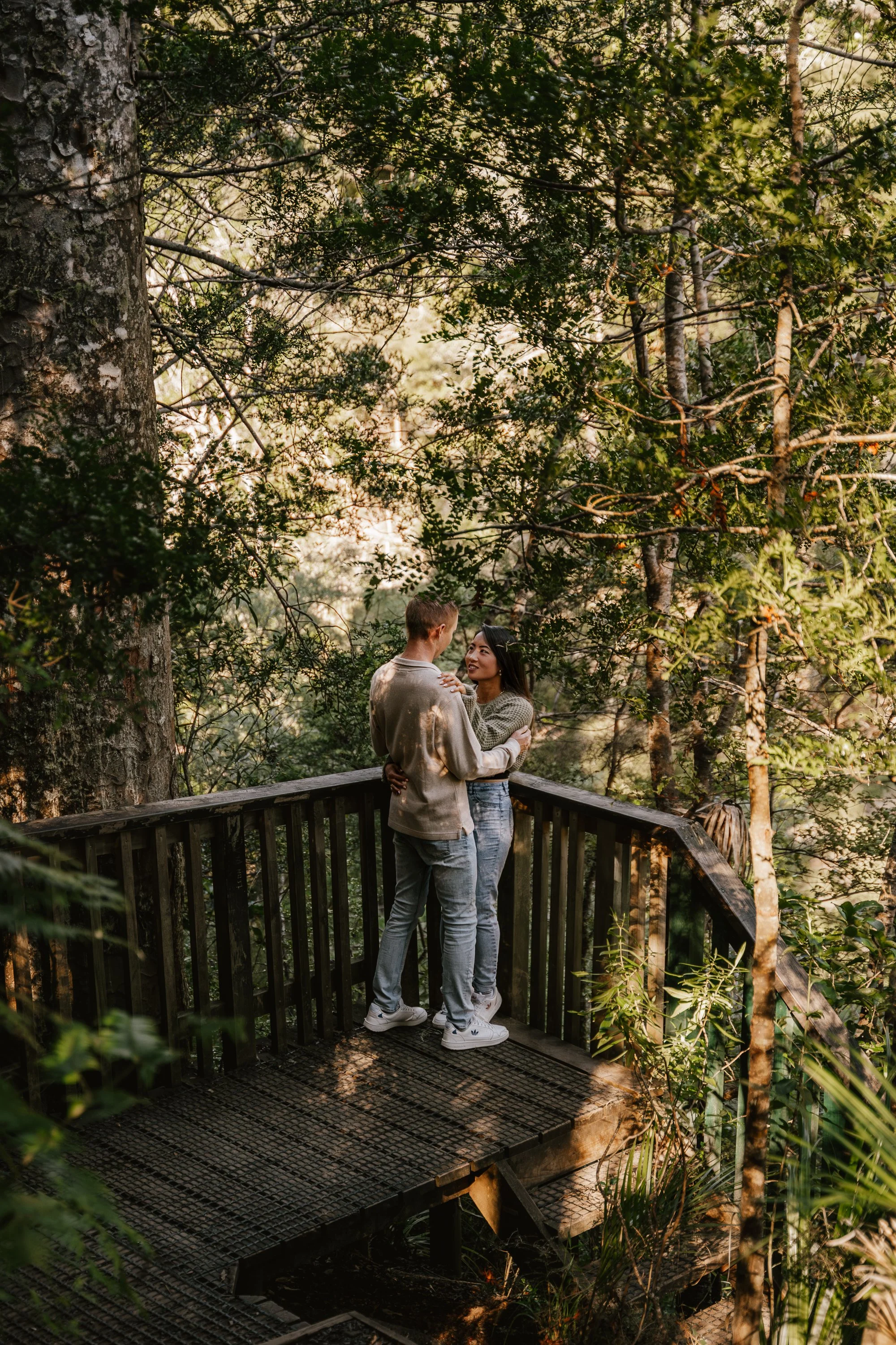 Two people, a man and a woman, standing close together on a wooden observation deck in a lush, green forest during daylight, smiling and looking at each other.