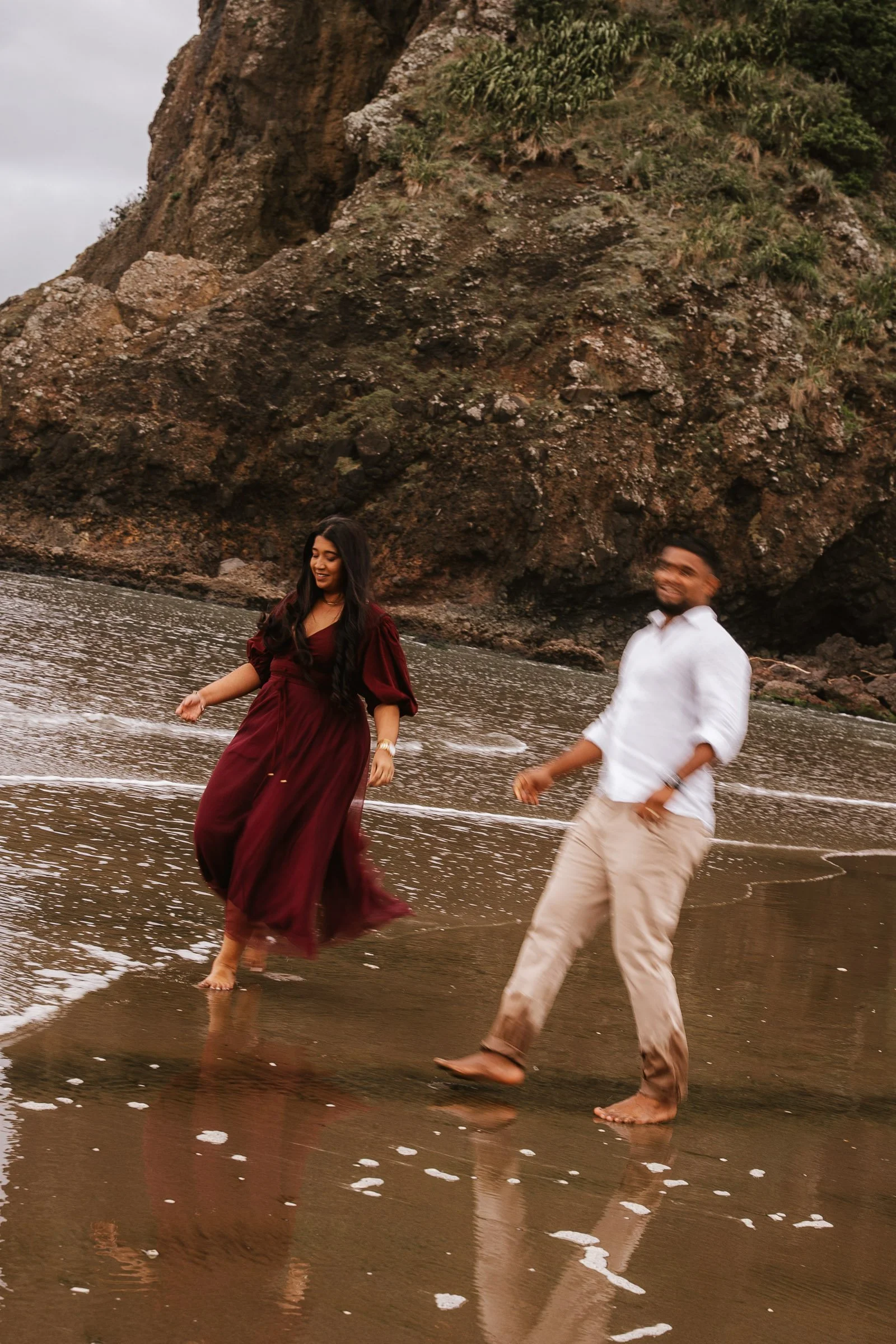 A man and woman walking barefoot on a beach, with waves at their feet. The woman is wearing a maroon dress, and the man is in a white shirt and beige pants. Behind them is a large rocky cliff with patches of greenery.
