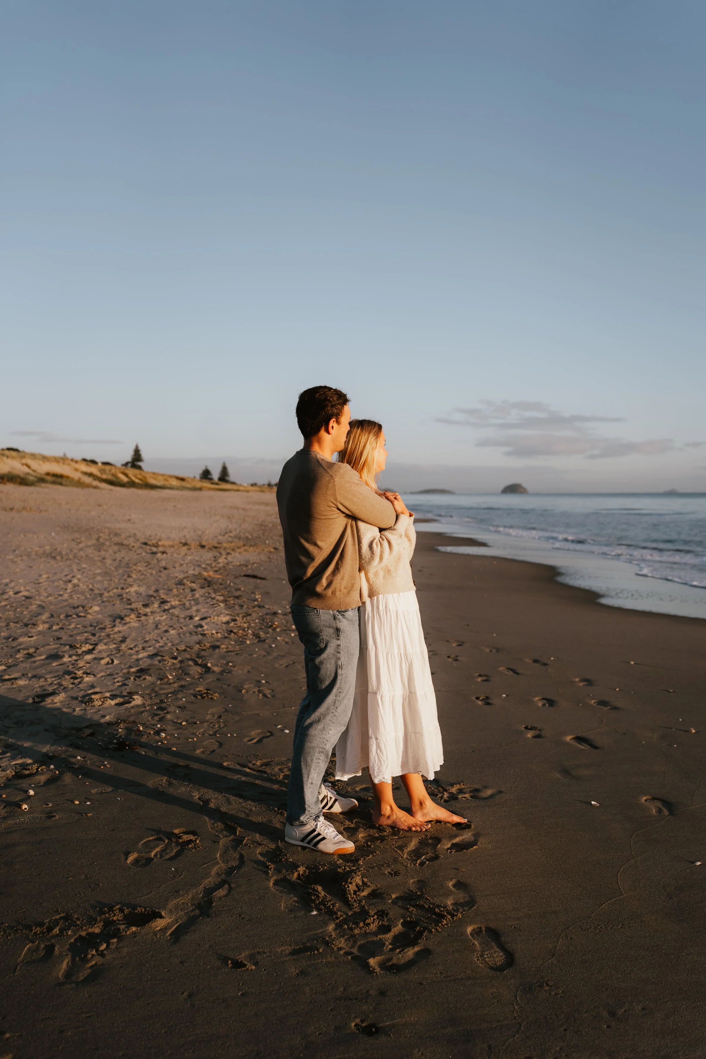 A man and woman stand on a beach at sunset, embracing and looking out at the ocean. The woman is barefoot in a white dress, while the man wears sneakers and jeans.