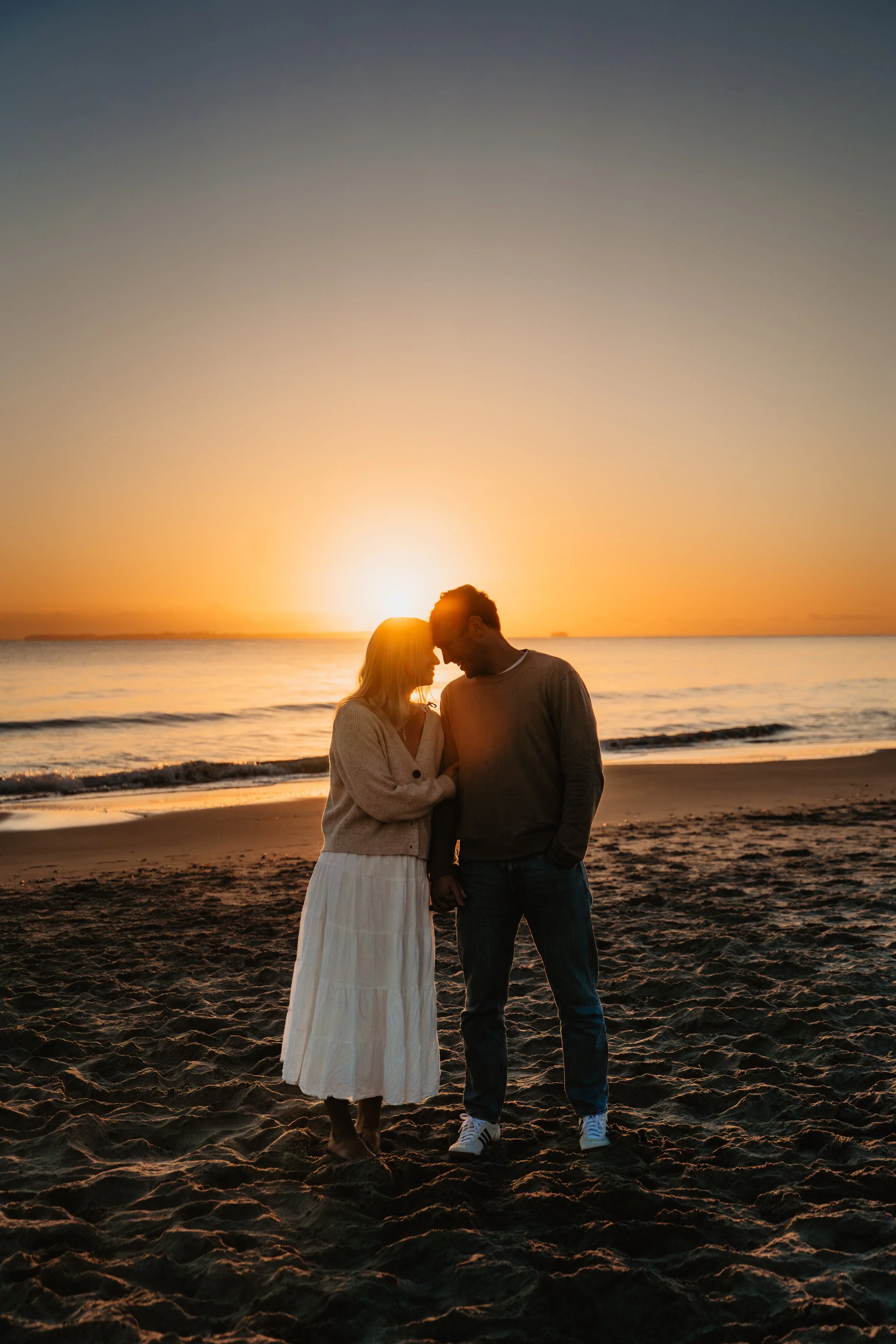 A couple standing on a beach at sunset, with their foreheads touching and holding hands, facing each other against the backdrop of the ocean and a setting sun.