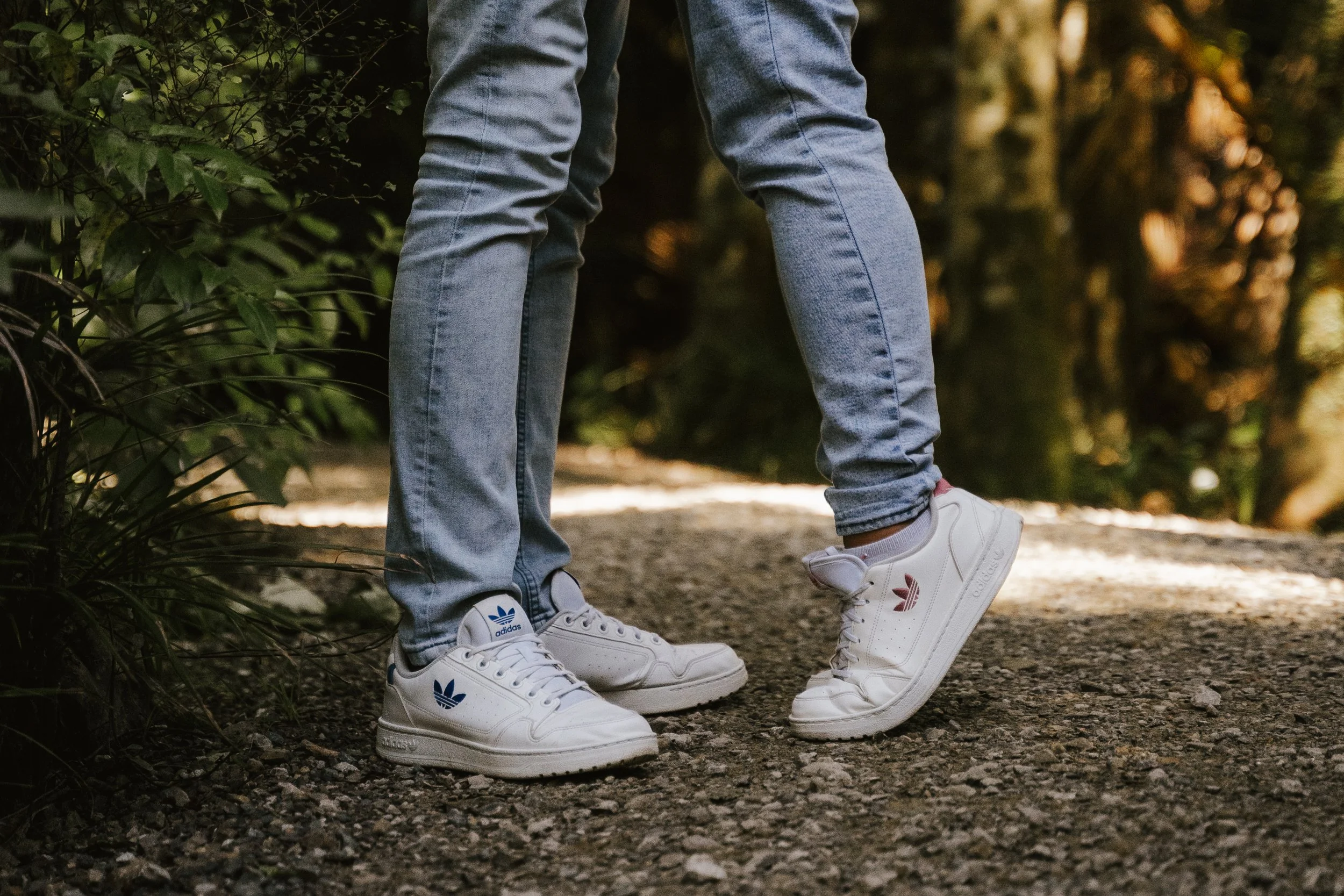 Close-up of two people standing on a dirt trail, wearing light blue jeans and white Adidas sneakers with blue and red logos, in a natural outdoor setting with greenery and trees.