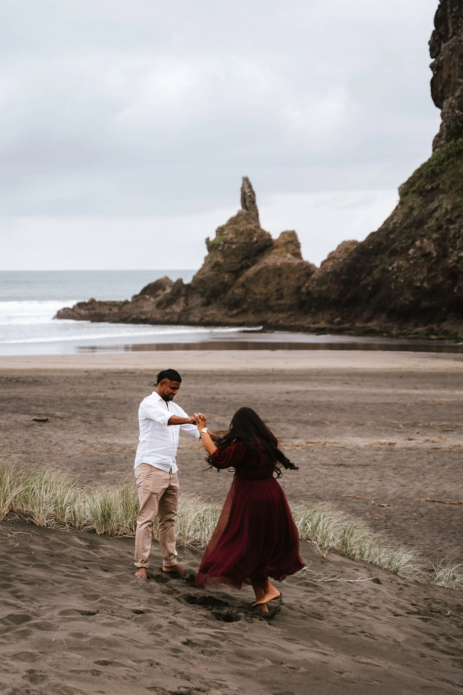 A man and woman are dancing on a beach with dark sand. The woman is wearing a long maroon dress, and the man is dressed in light-colored pants and a white shirt. They are holding hands and smiling. In the background, there are large rocks and an overcast sky.
