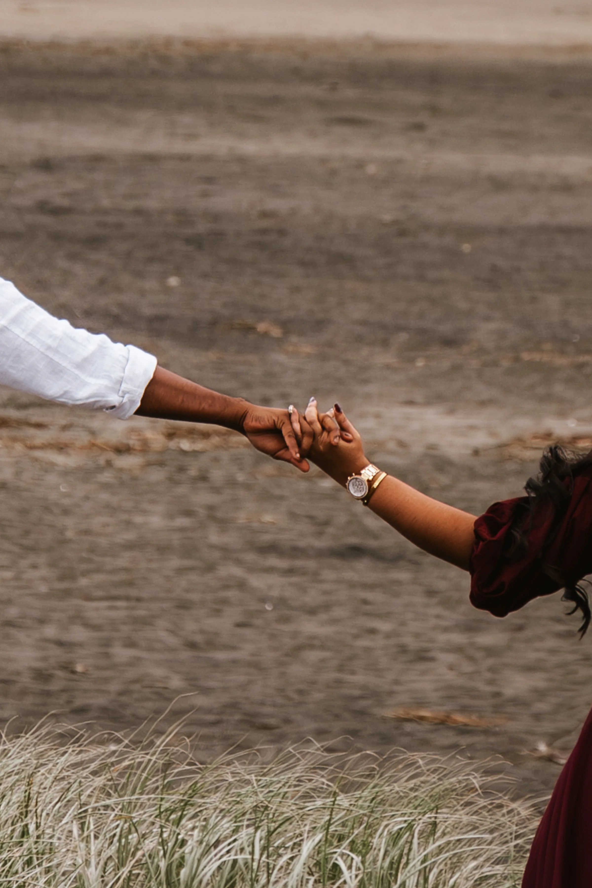 A person wearing a white shirt holding hands with another person wearing a wristwatch in a scenic outdoor setting with tall grasses and a sandy landscape.