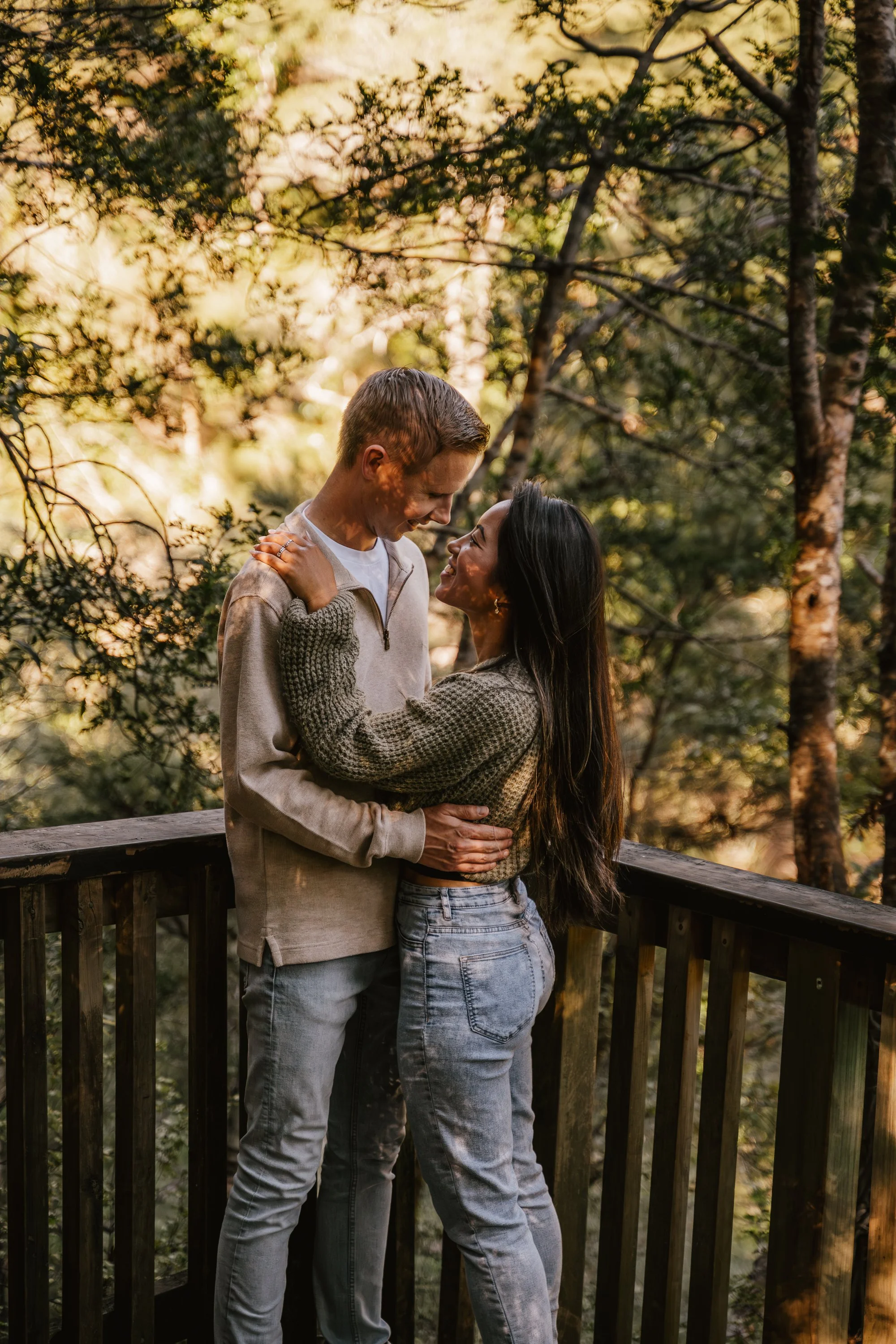 A couple standing on a wooden deck outdoors during autumn, smiling and looking at each other while embracing with trees and foliage in the background.