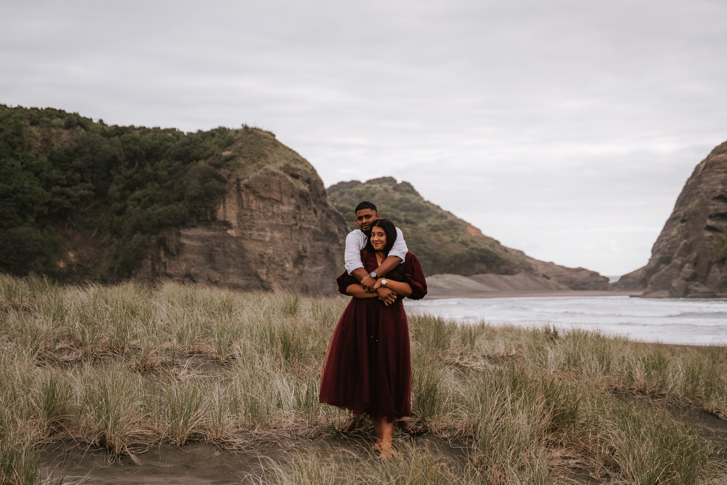 A couple standing on a grassy beach with large cliffs and cloudy sky in the background. The woman in front is wearing a long burgundy dress, and the man behind her is embracing her with his arms around her shoulders.