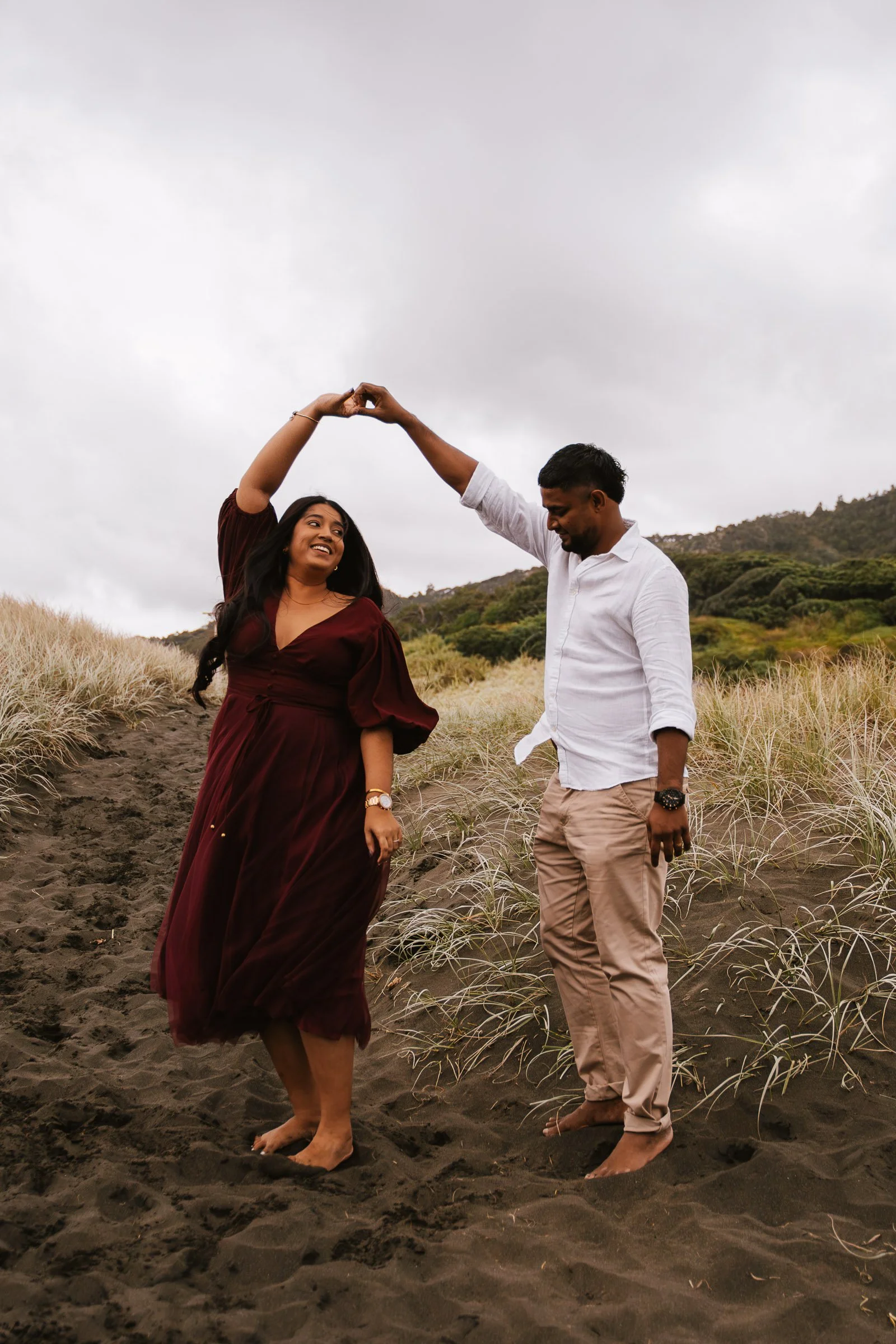 A couple dancing barefoot on a sandy path surrounded by grass, with overcast sky and hilly landscape in the background.