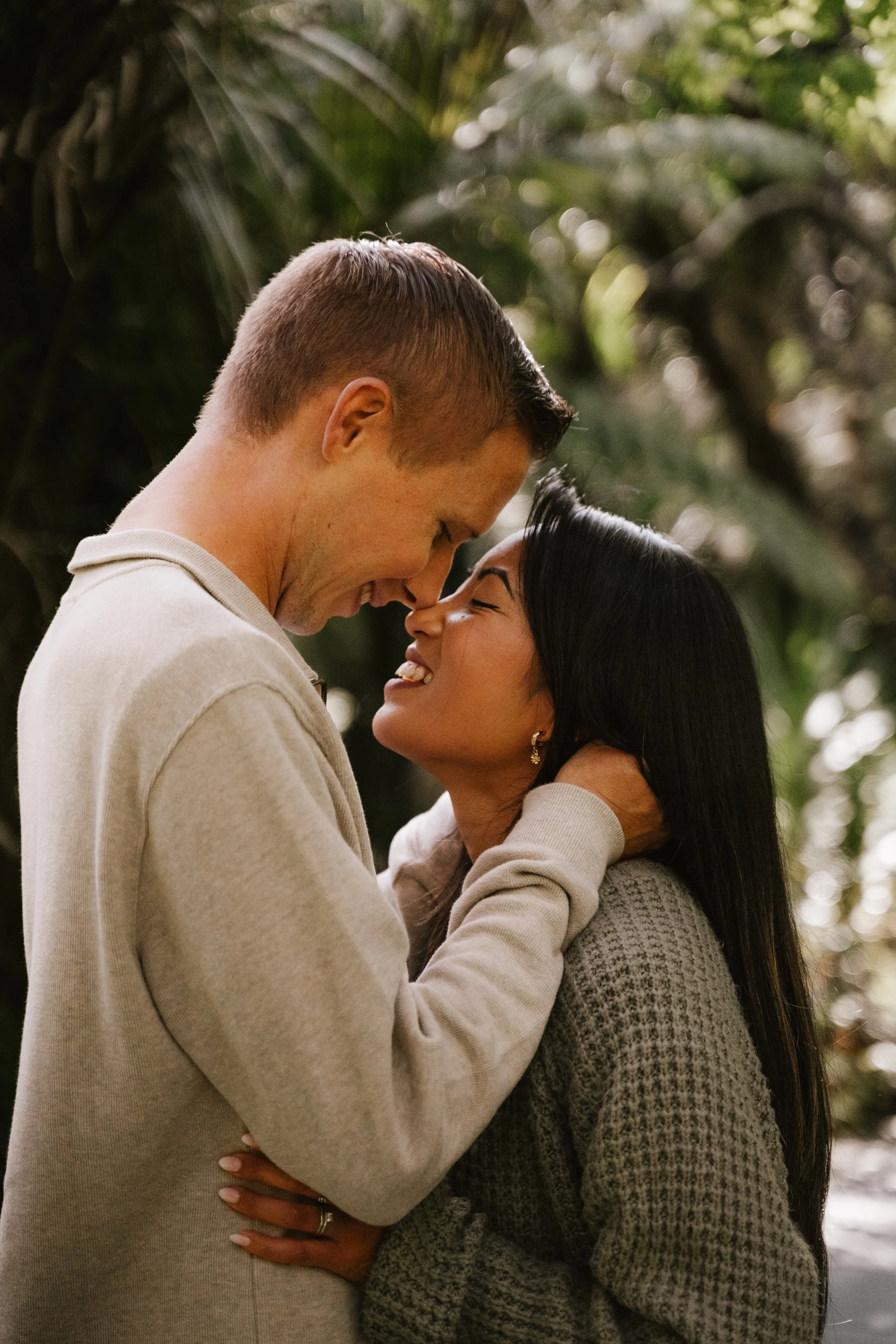 A couple is smiling and touching foreheads, in an outdoor setting with lush green foliage in the background.