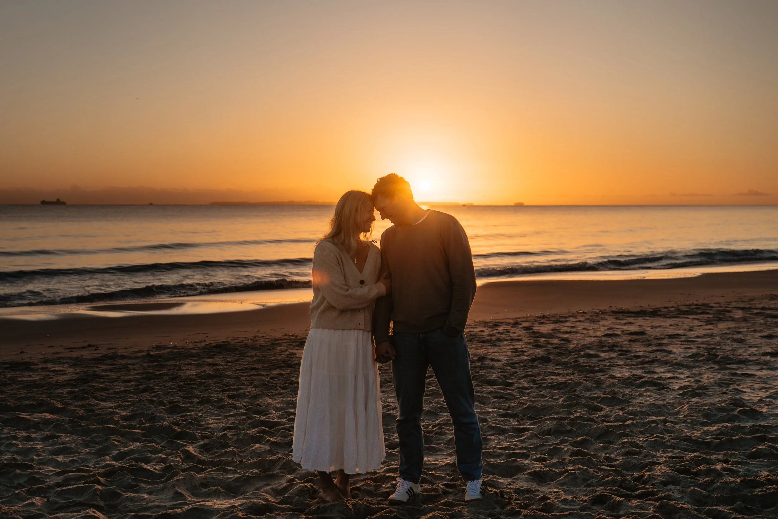 A couple standing on a beach at sunset, with their foreheads touching and smiling at each other.