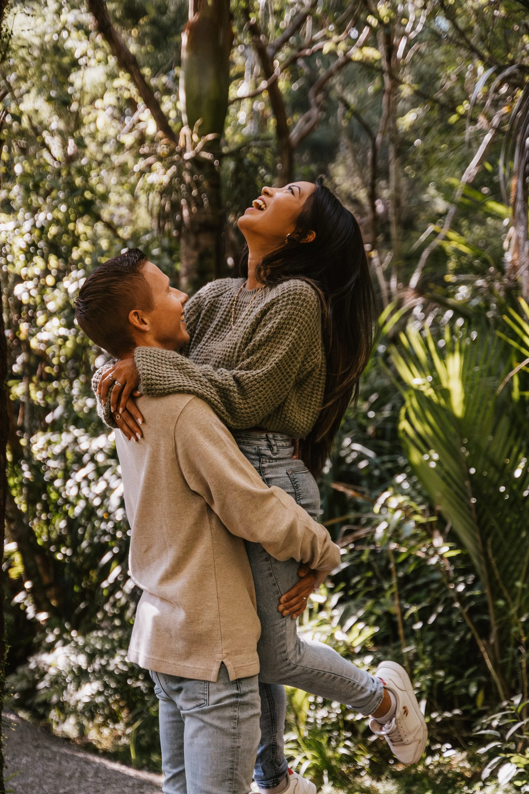 A man lifting a woman in a joyful embrace outdoors among trees and plants, both smiling and laughing.