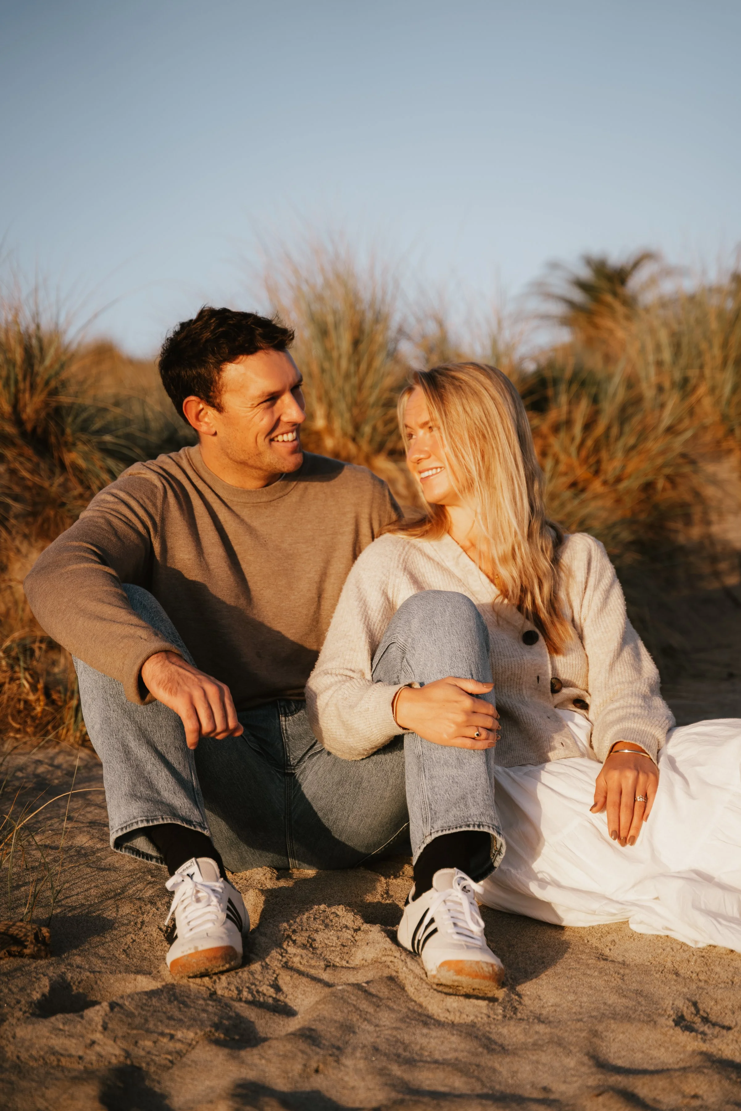 A smiling man and woman sitting on a sandy beach near tall grass during sunset, looking at each other and enjoying the moment.