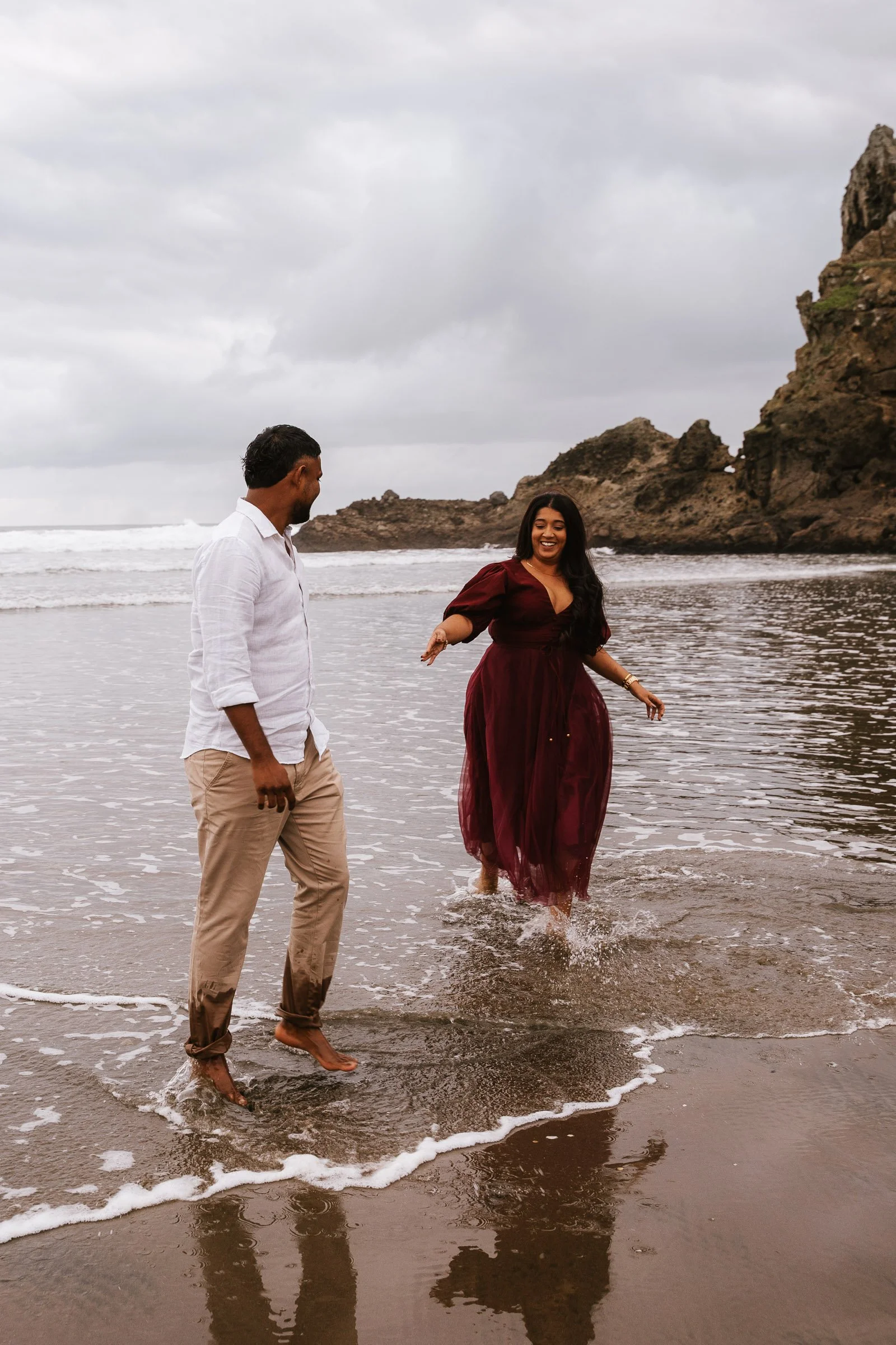 A man and woman walking barefoot in the ocean waves at the beach on a cloudy day. The man is wearing a white shirt and beige pants, and the woman is wearing a maroon dress, smiling and splashing water.