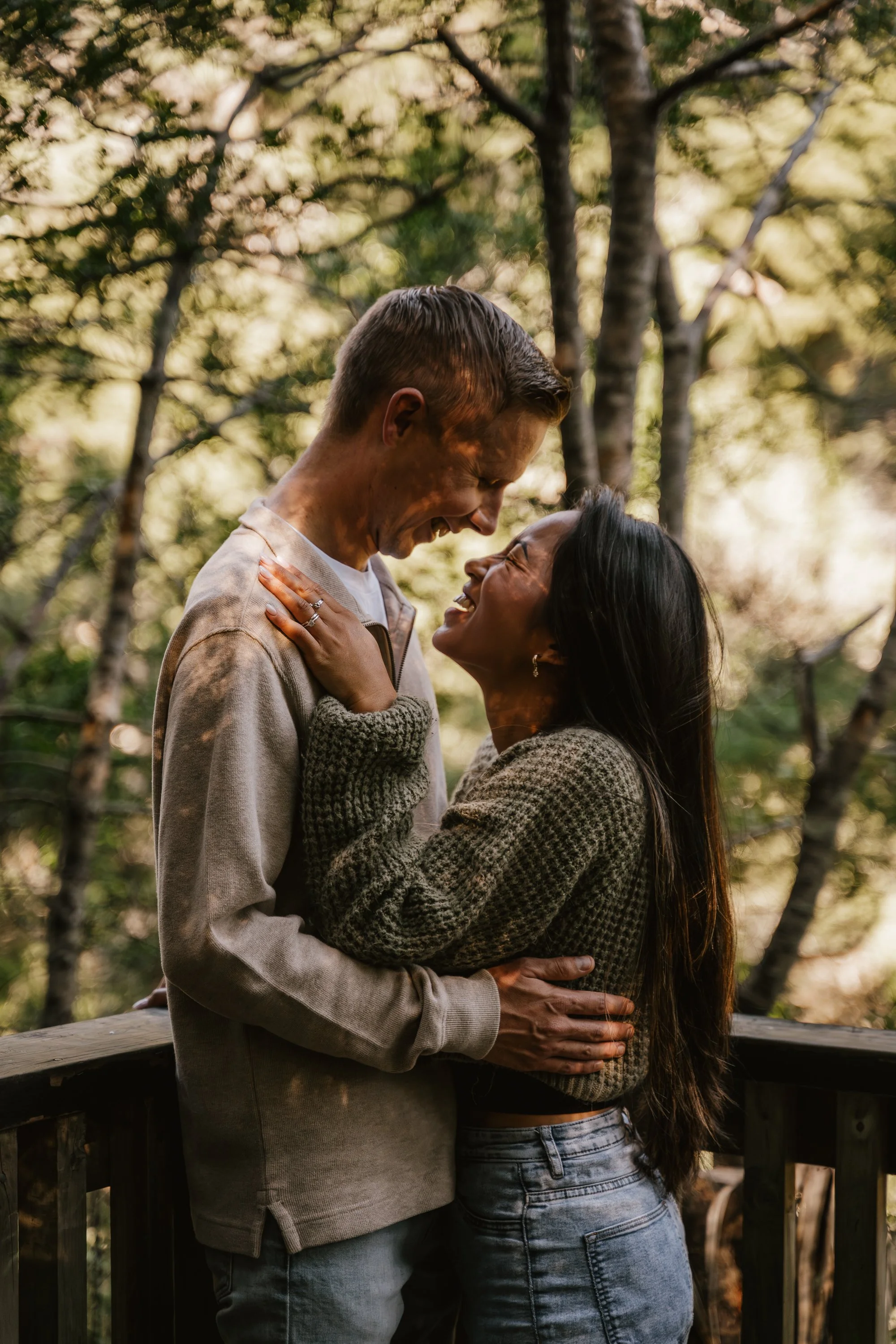 A couple sharing an affectionate moment outdoors in a wooded area, with the man smiling and the woman laughing, both holding each other.