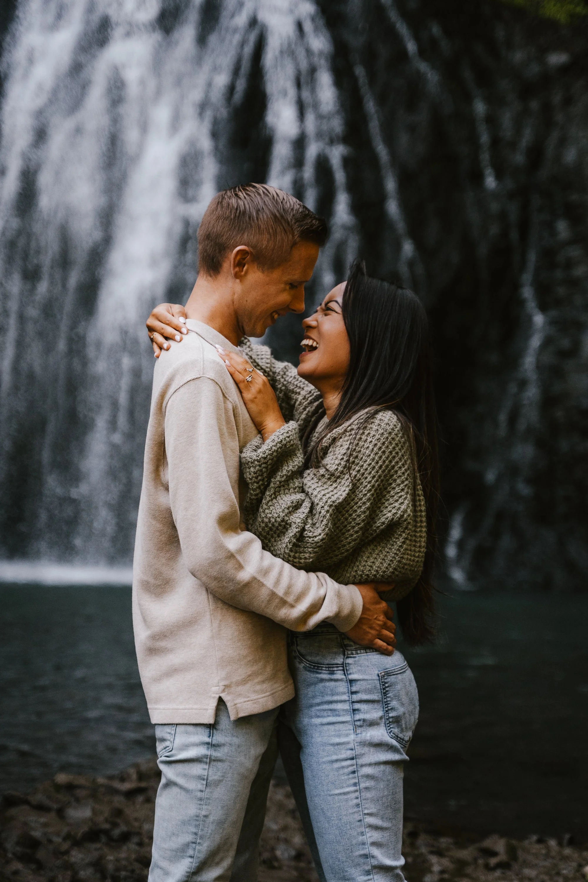 A couple standing close together in front of a waterfall, smiling and looking into each other's eyes.