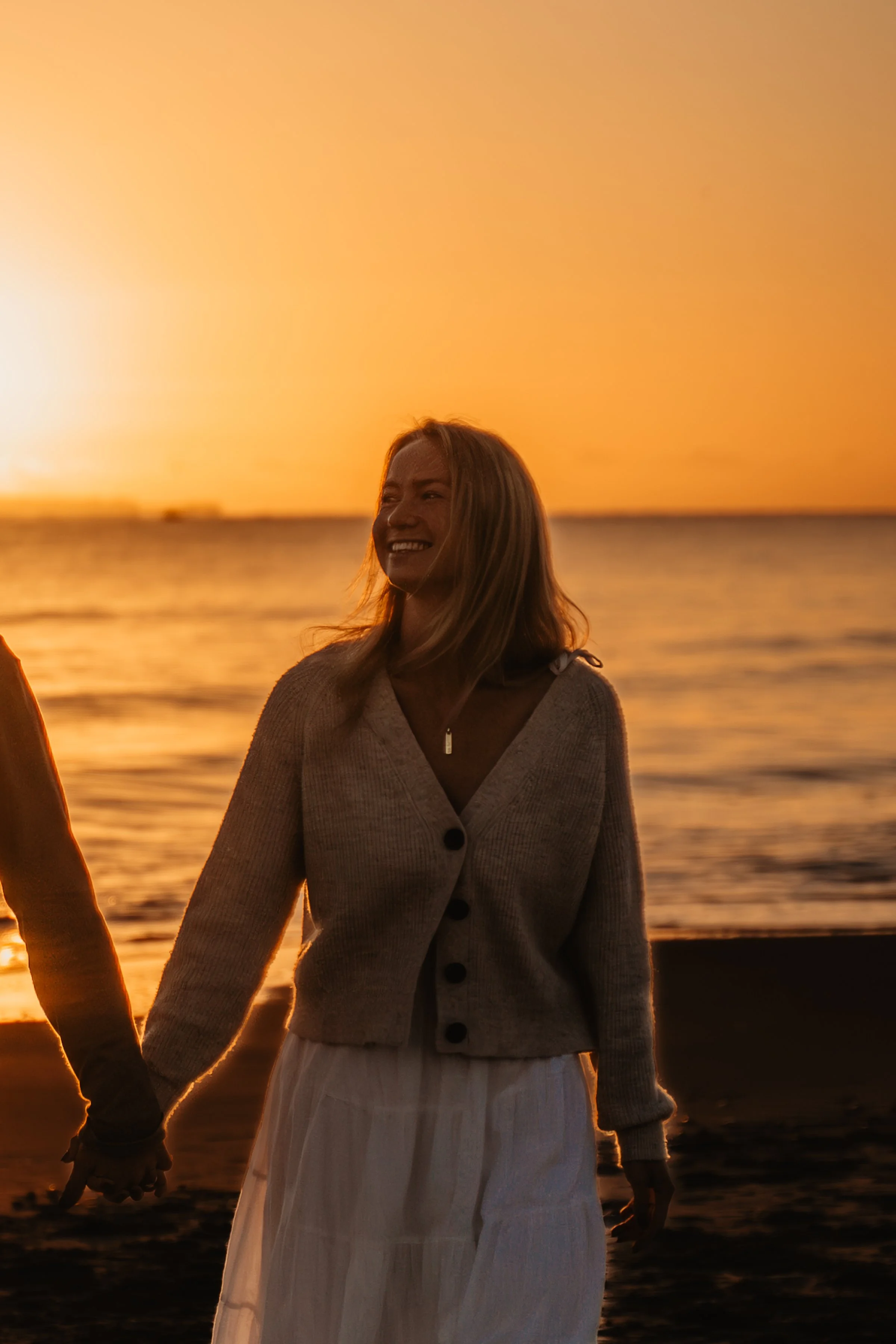 A woman with blonde hair smiling and holding hands with someone at sunset on a beach, with the ocean and sky in the background.