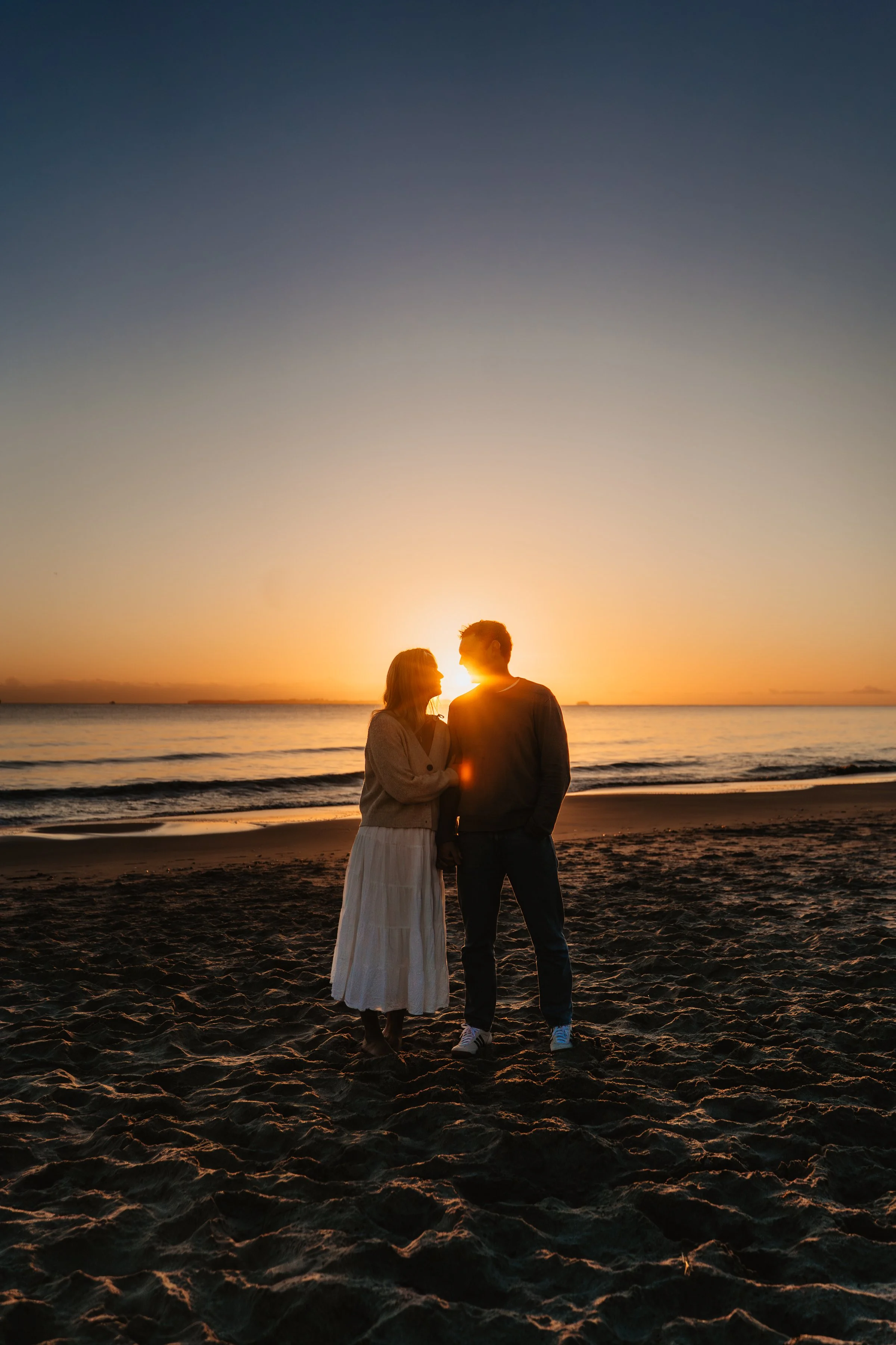 A couple standing on the beach at sunset, holding hands and gazing at each other, with the ocean and sky in the background.