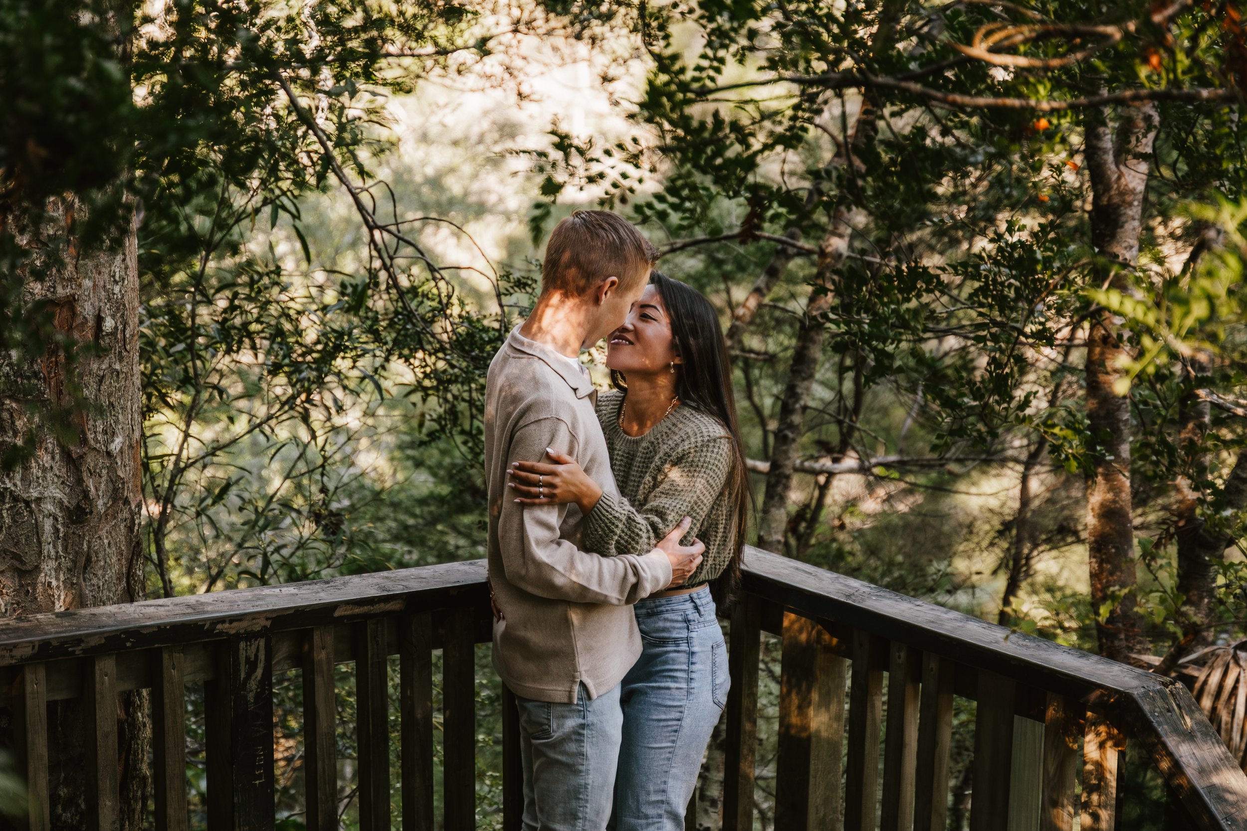 A couple embraces on a wooden bridge in a forest, smiling at each other with trees and sunlight in the background.