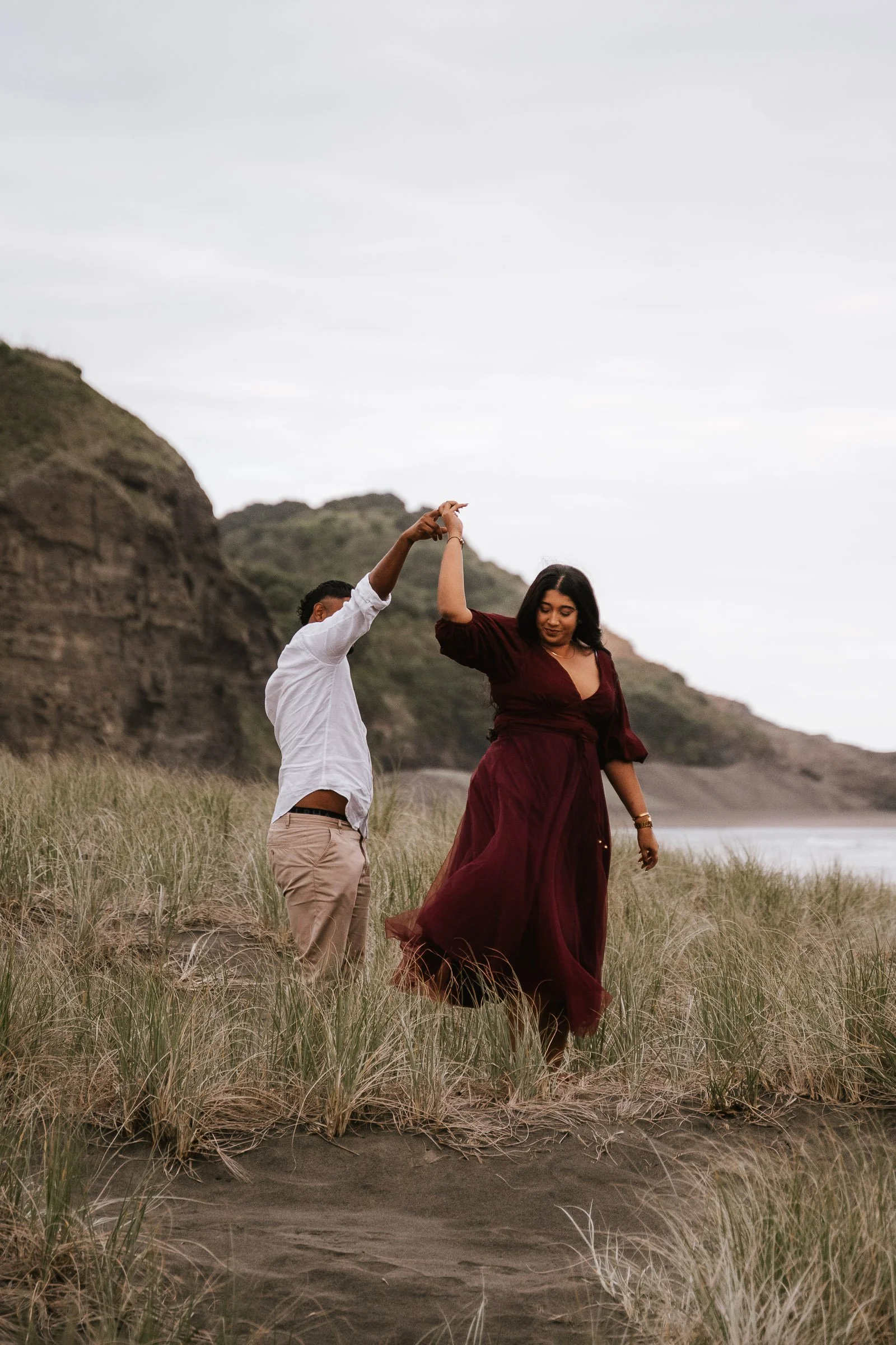 A couple dancing on a grassy beach, with rocky cliffs and overcast sky in the background.