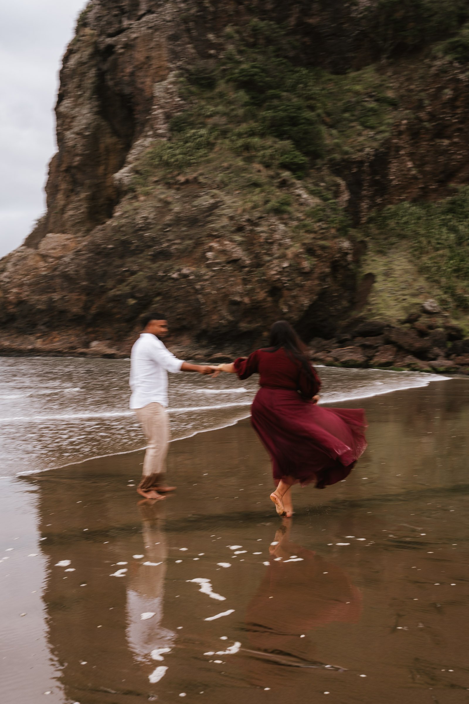 A couple holding hands and dancing on the wet sandy shore at the beach, with rugged rocky cliffs covered in greenery in the background.